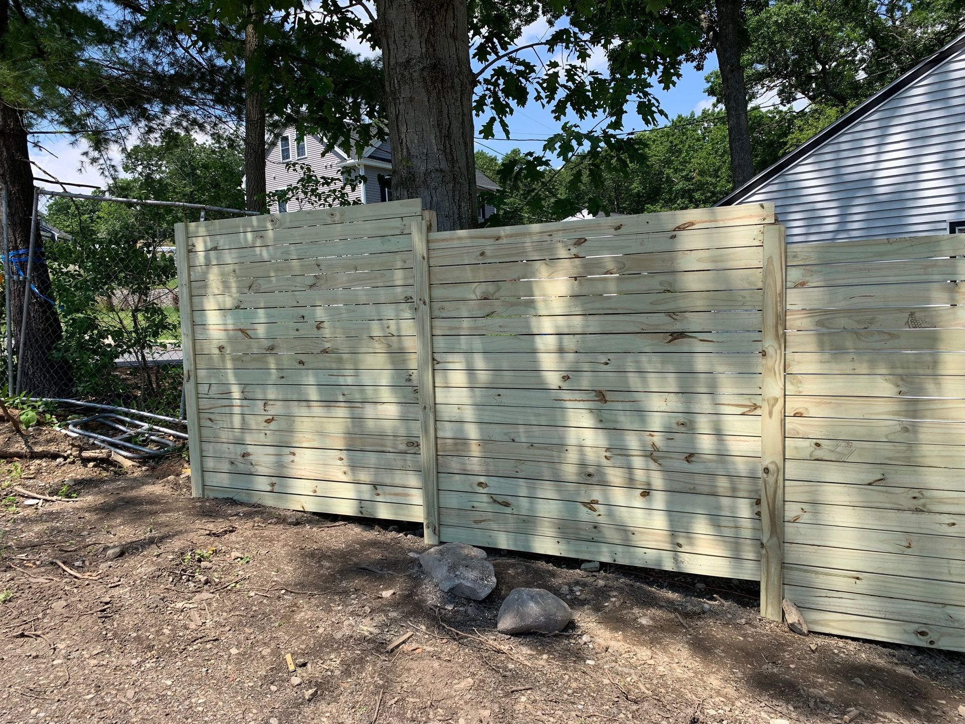 Wooden fence in front of trees and a house on a sunny day.