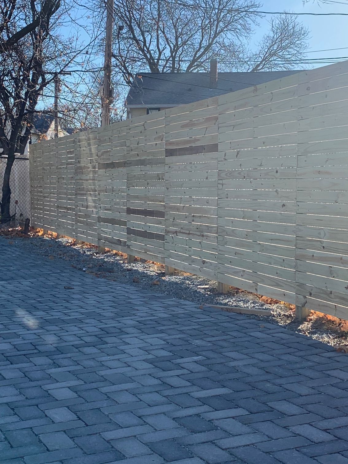 Gray brick driveway with a light-colored horizontal slatted wooden fence, leafless trees, and a house in the background.