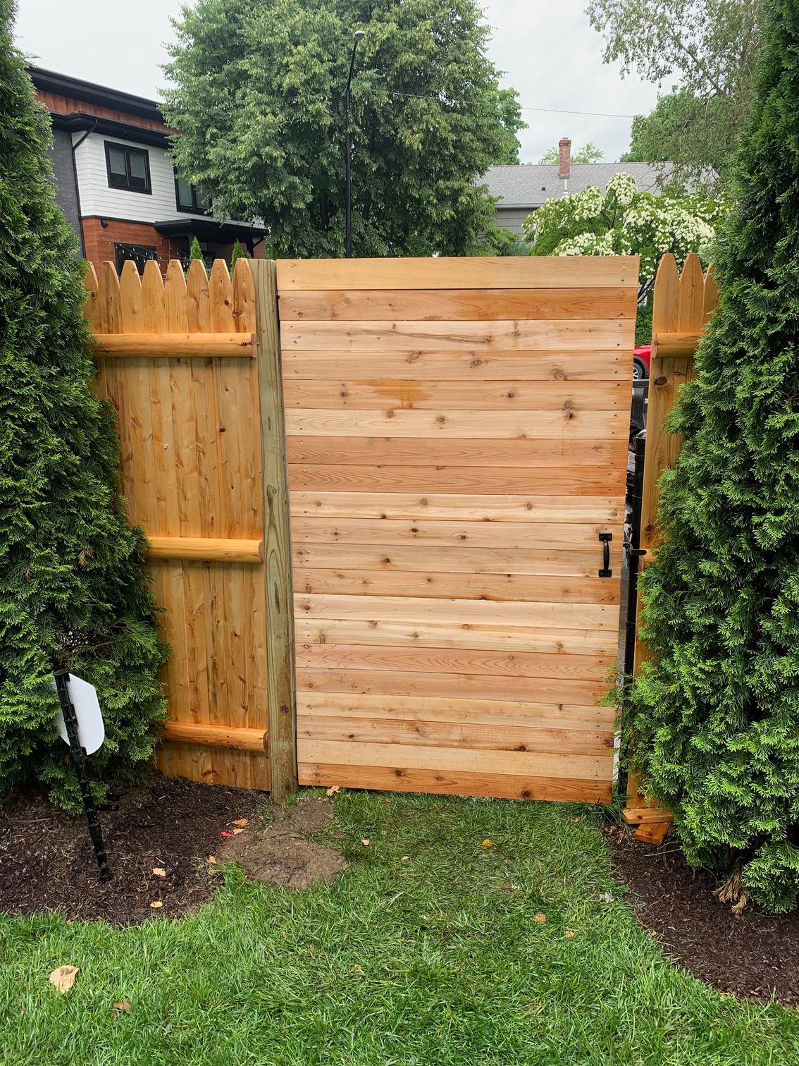 Wooden gate in a cedar fence, flanked by evergreen trees. The gate is closed, in a grassy yard.