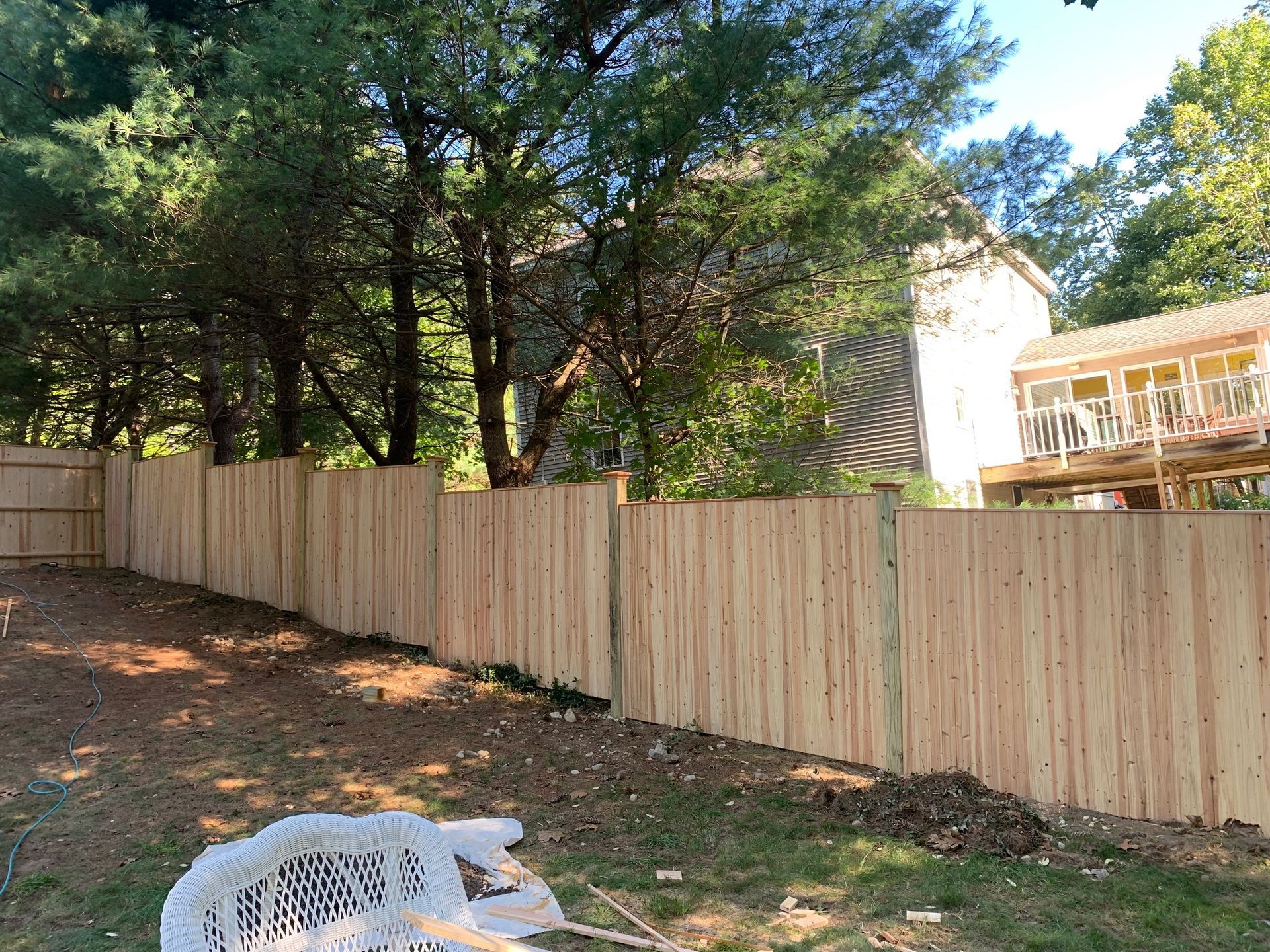 Wooden fence in a backyard, trees, house in background. Sunny day, construction debris.