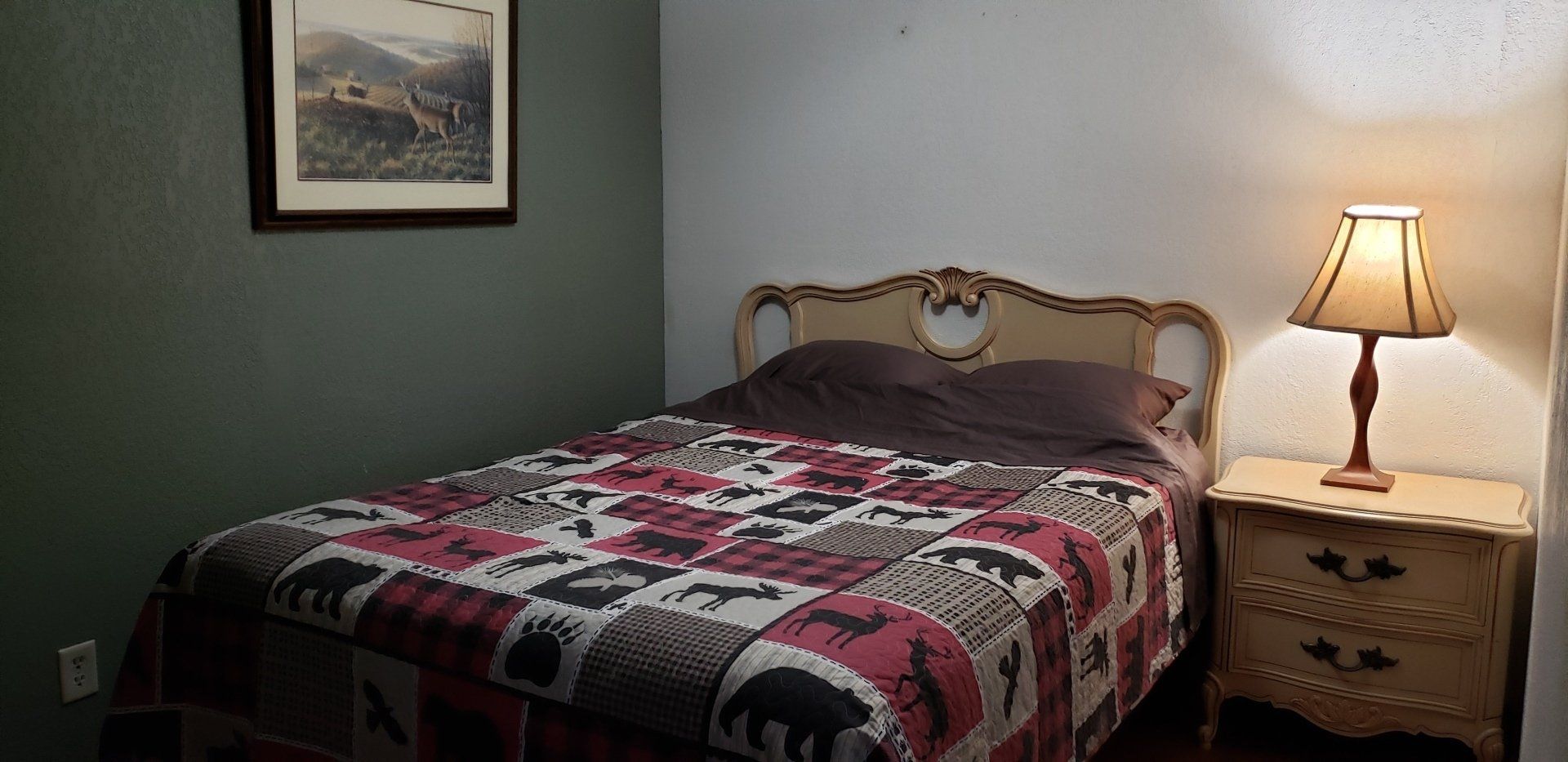A bedroom with a bed , nightstand , lamp and picture on the wall.