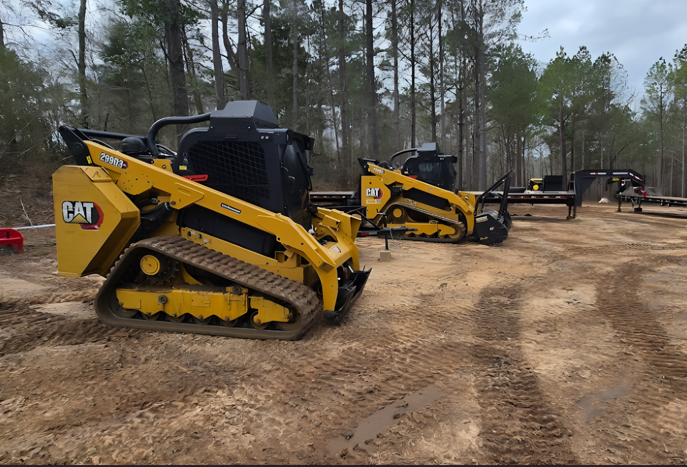 Two yellow Caterpillar skid steer loaders on a dirt lot near trees, ready for work.