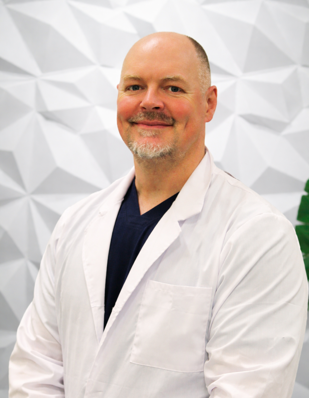 A professional in a white medical coat and dark blue scrubs smiles in front of a white geometric textured background.