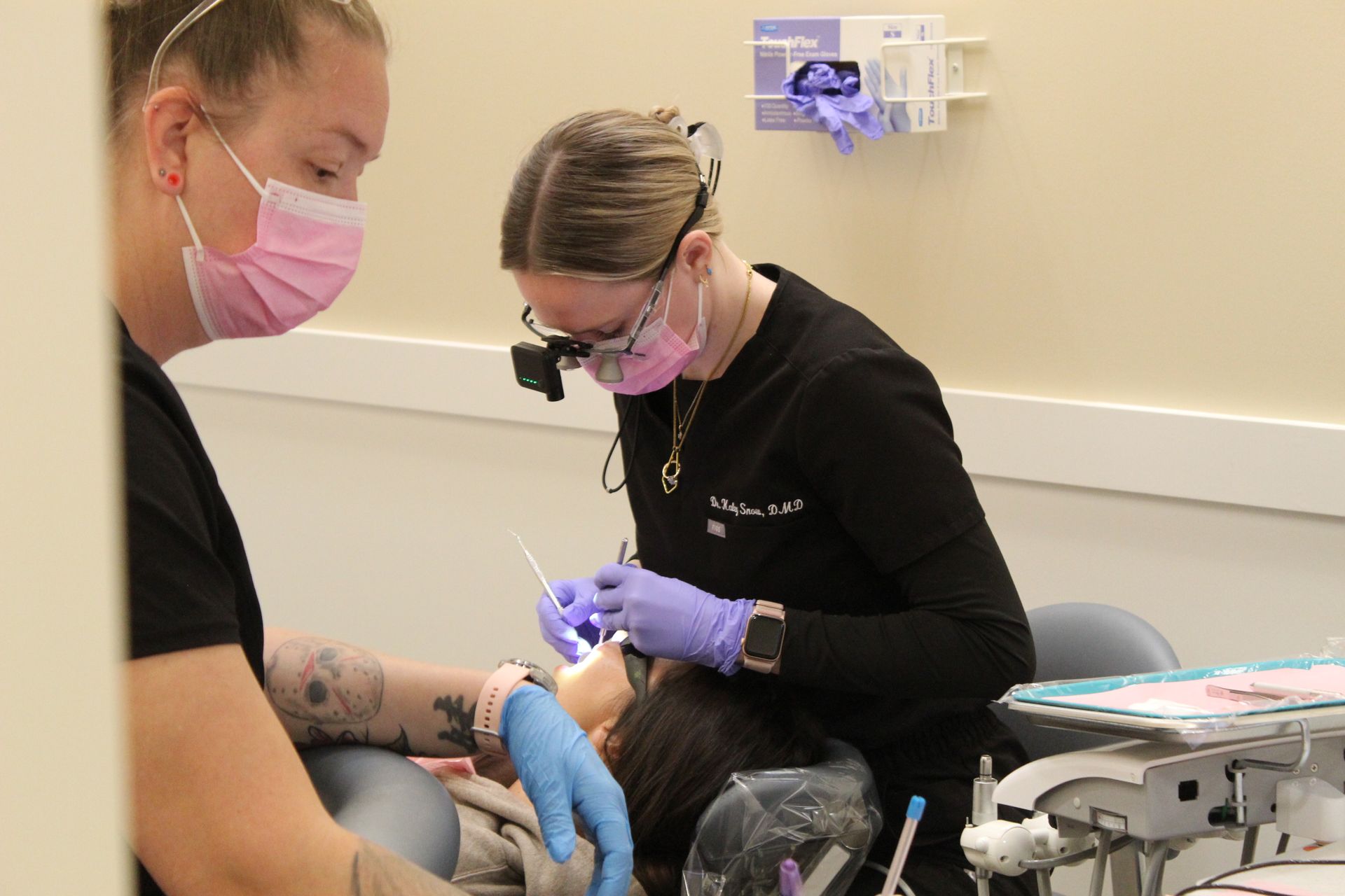 Dentist examining patient's teeth in a dental office. Another assistant nearby. Both wearing masks and gloves.
