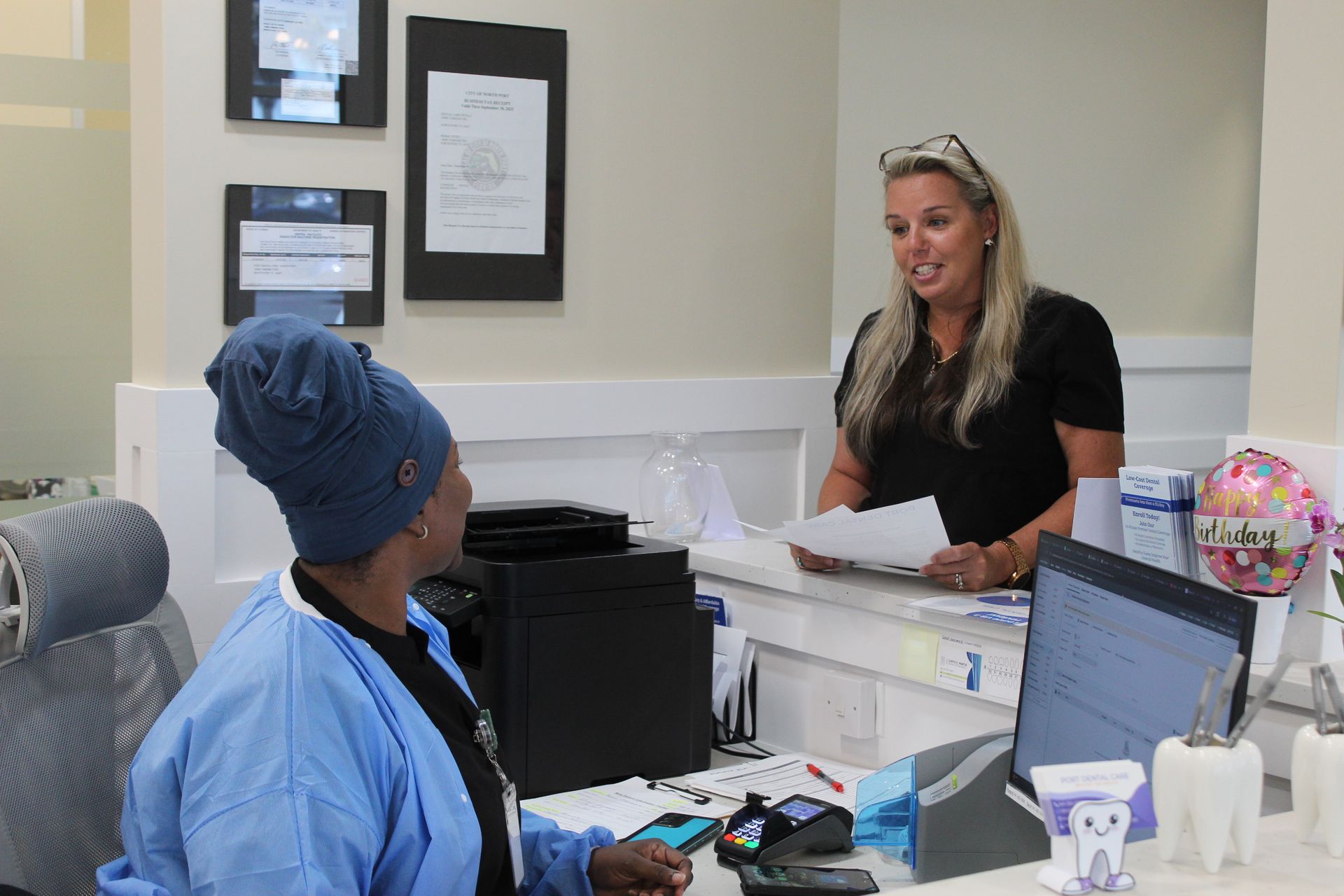 Two women at a dental office reception desk. One hands papers to the other while smiling.