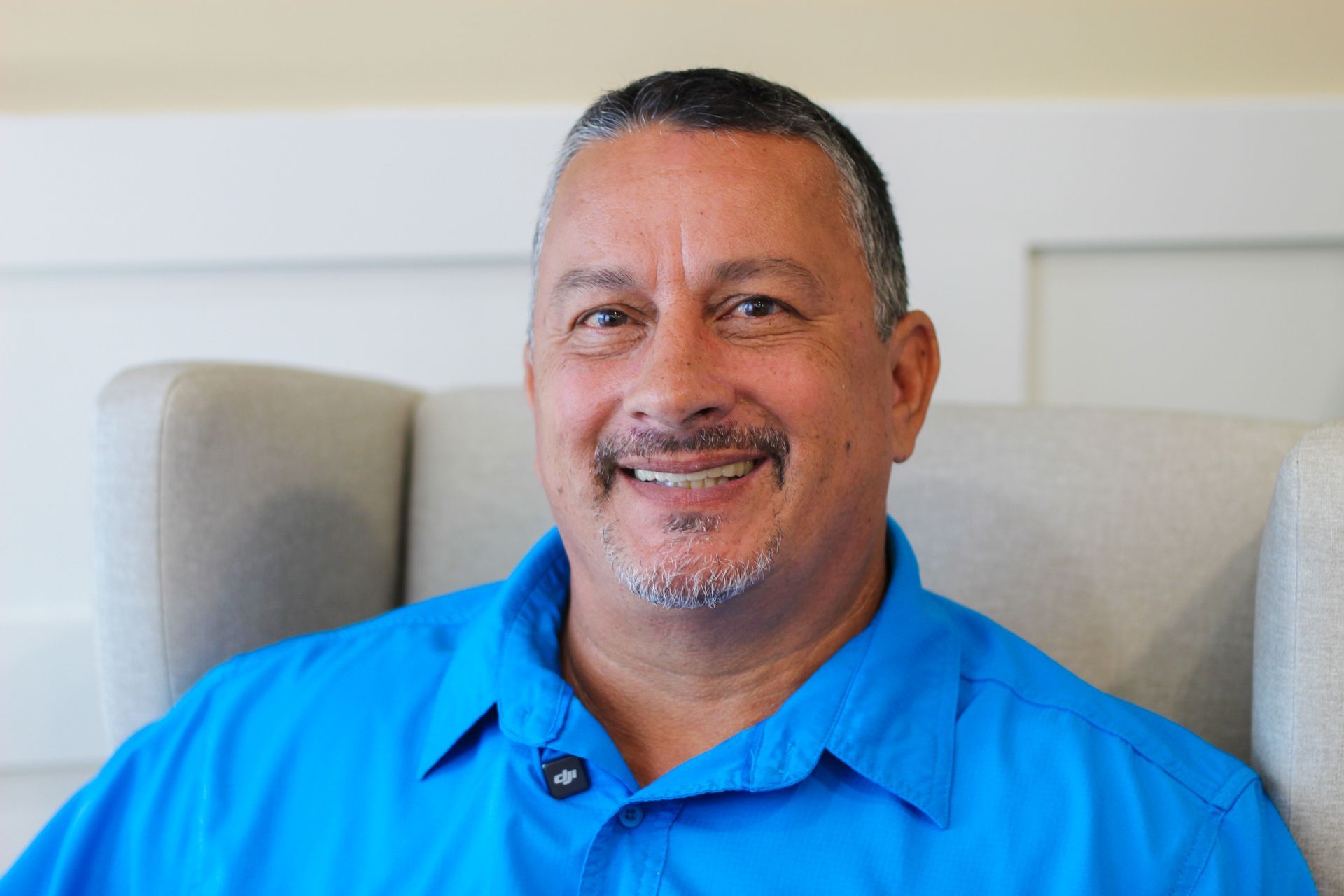 Man in a blue shirt smiles at the camera, seated in a light gray chair.