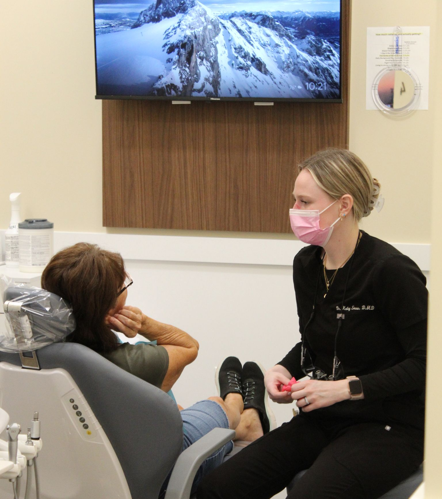 Dentist in mask consults with a patient in dental chair, looking at a model of teeth in hand.