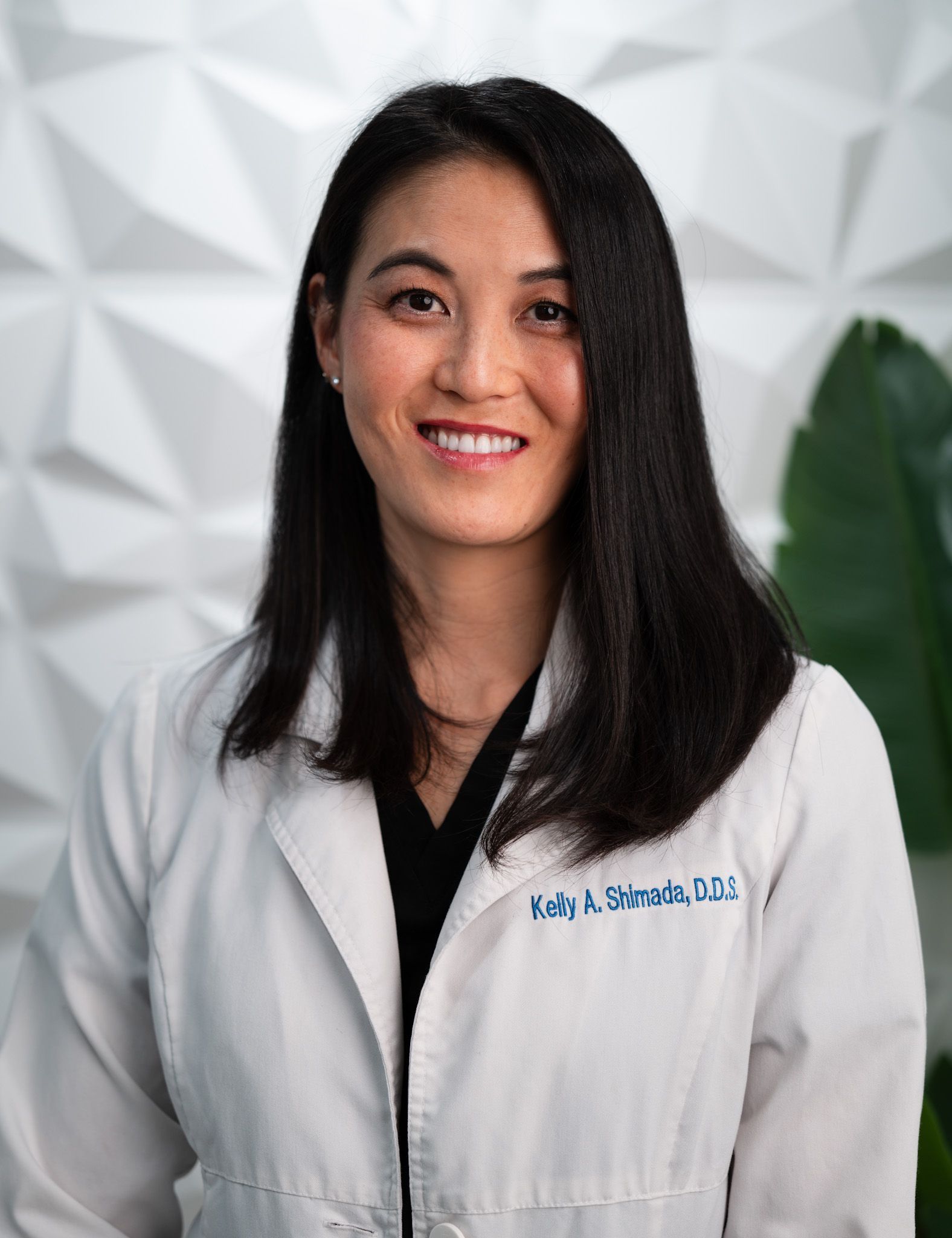 Woman in white coat, smiling, against a textured white wall.