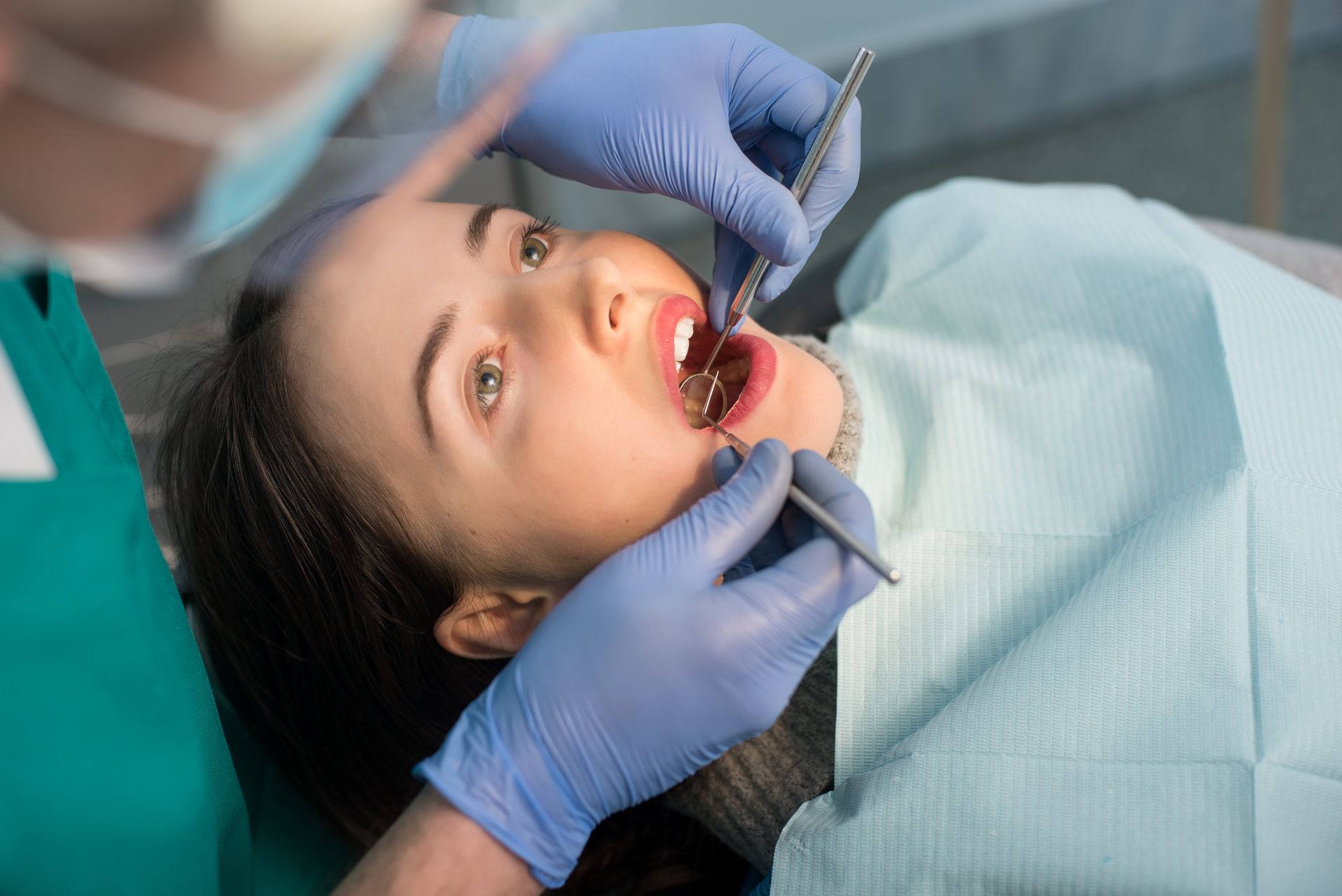 A dentist in blue gloves uses dental tools to examine a patient’s open mouth during a clinical checkup.