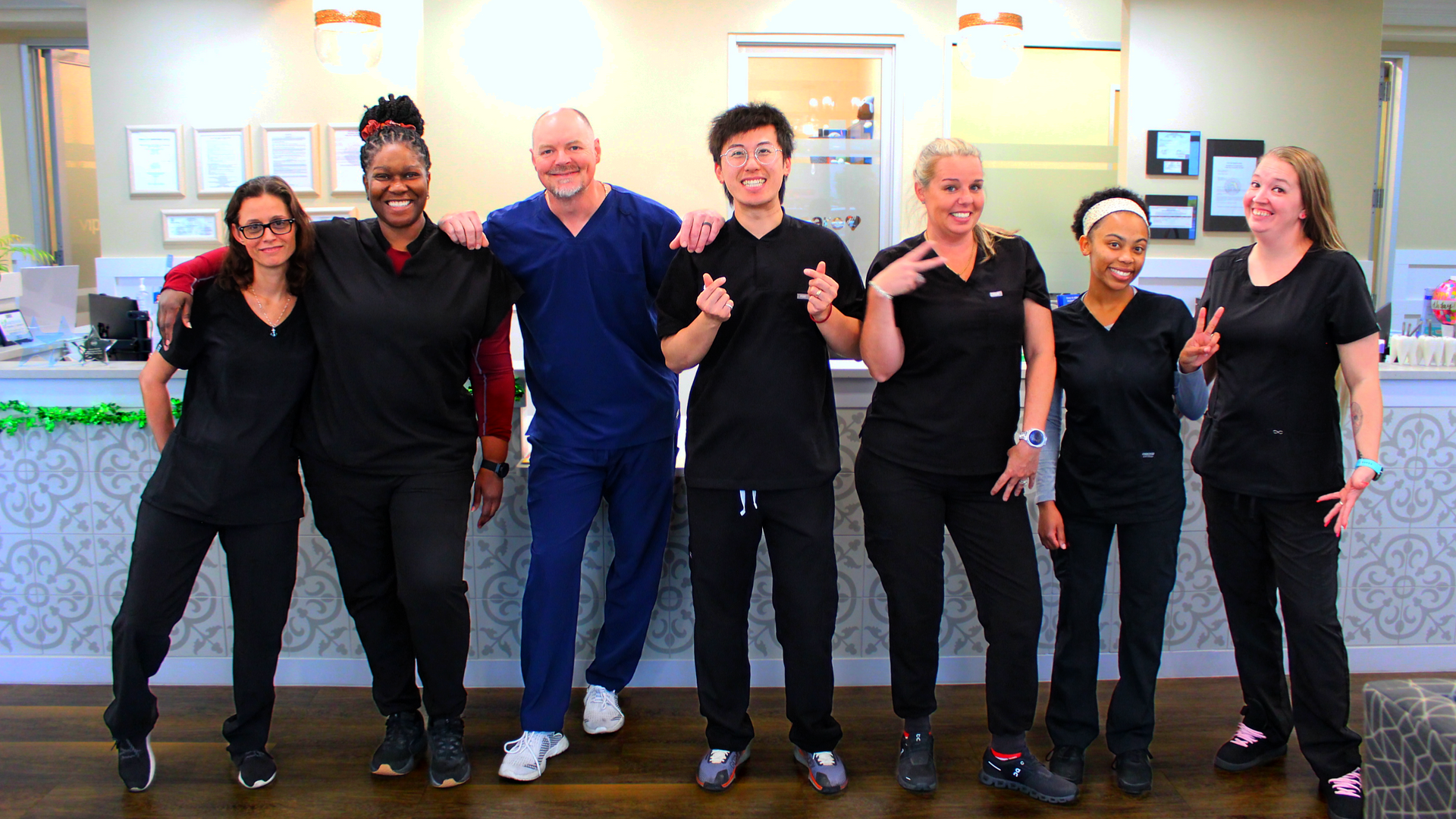 A group of seven dental office staff members in black scrubs and one in blue scrubs stand in a row smiling at a front desk.
