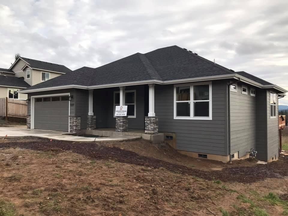 Gray two-story house with a wooden deck, surrounded by green grass and trees under a blue sky.