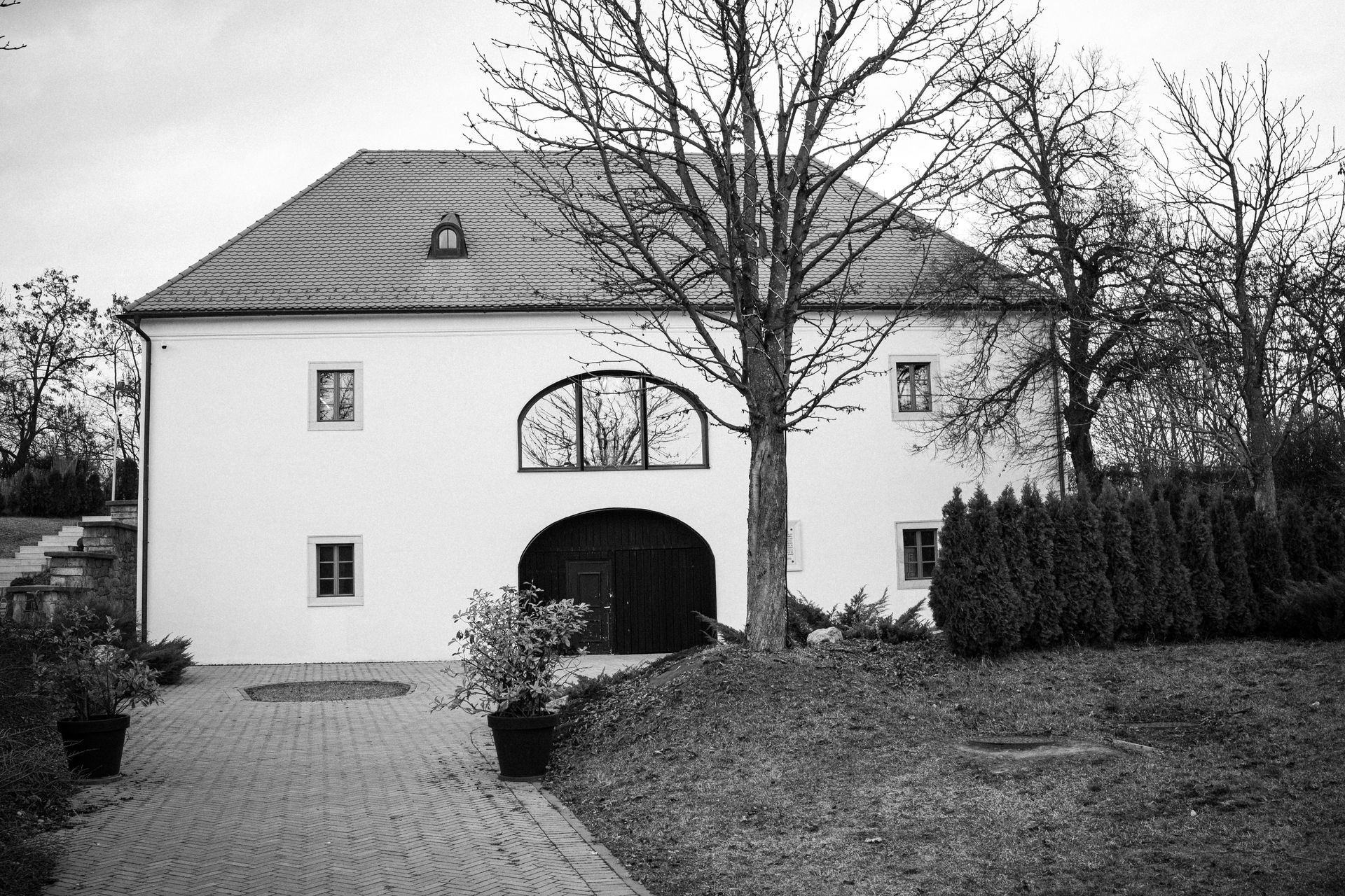 Black and white photo of a white building with a large arched doorway, brick path, and trees.