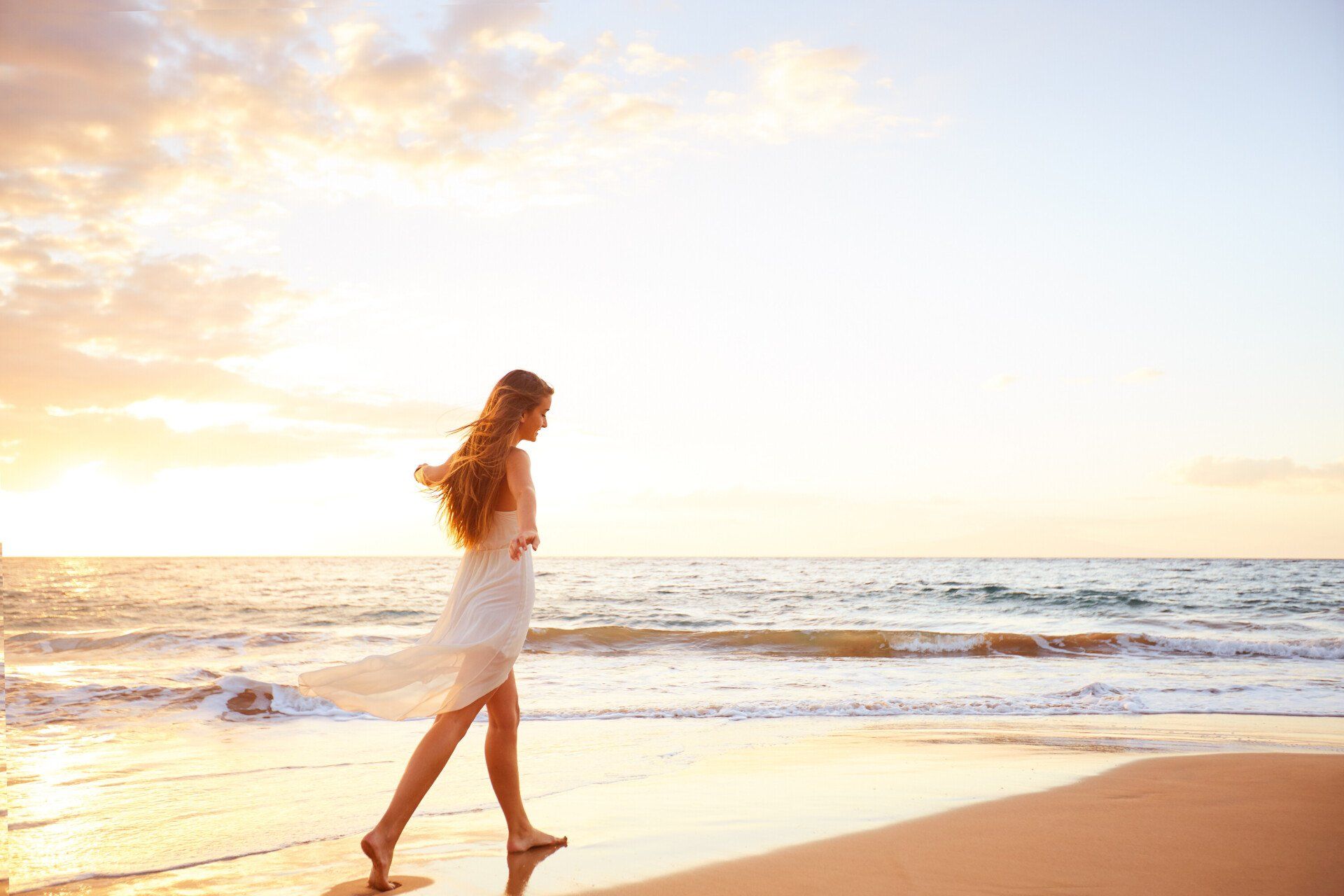 A woman in a white dress is walking on the beach at sunset.