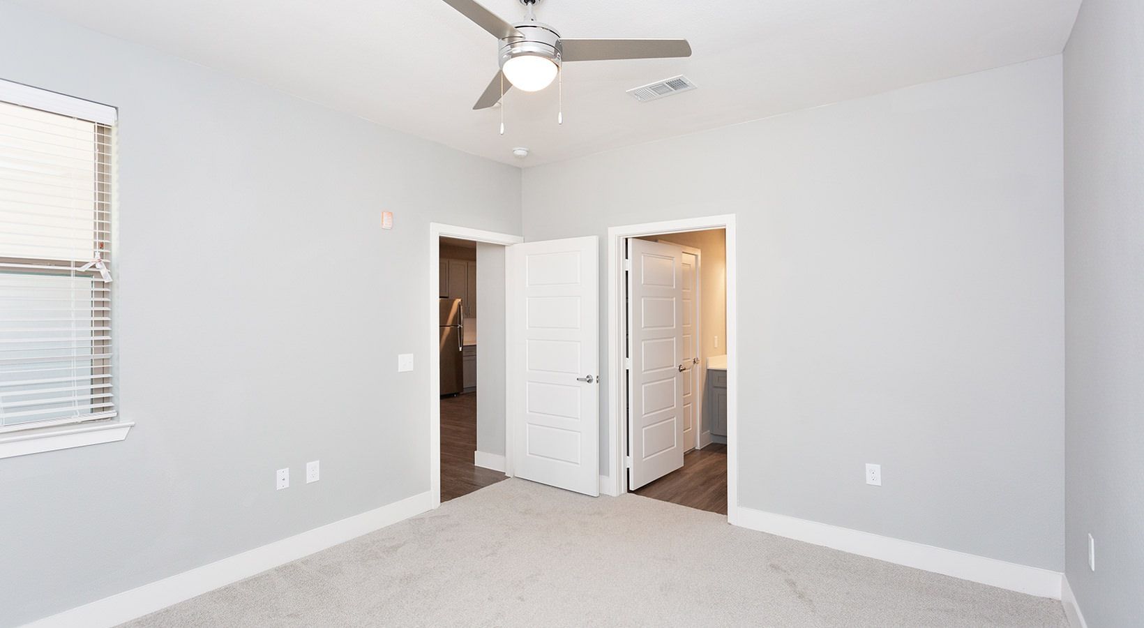 Empty bedroom with gray walls, white trim, ceiling fan, window blinds, and two doors to kitchen and bathroom.