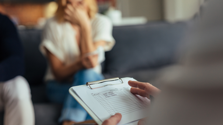 A man is holding a clipboard in front of a woman sitting on a couch.