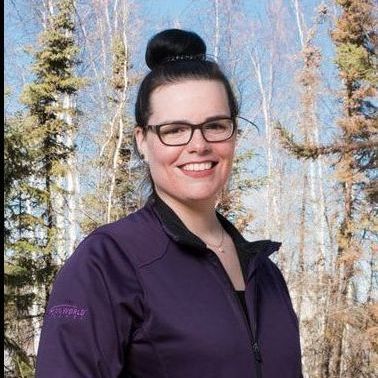 A woman wearing glasses and a purple jacket is smiling in front of trees.