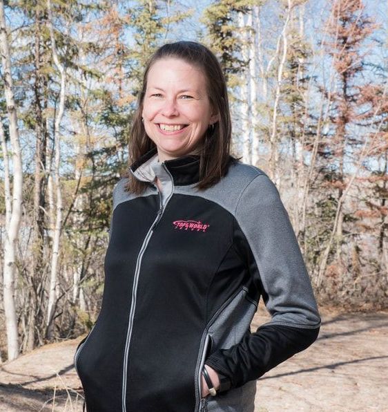 A woman wearing a black and grey jacket is smiling in front of trees