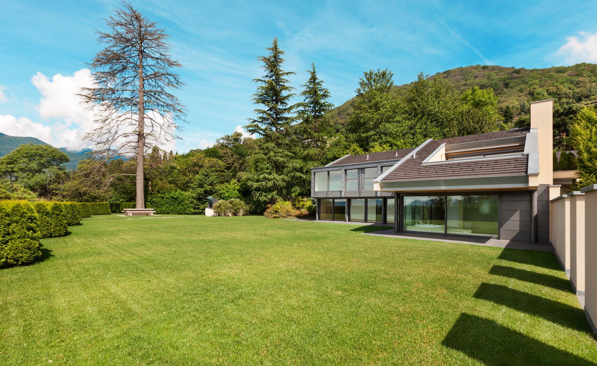 Sunny backyard lawn beside a modern house with trees and green hills in the background