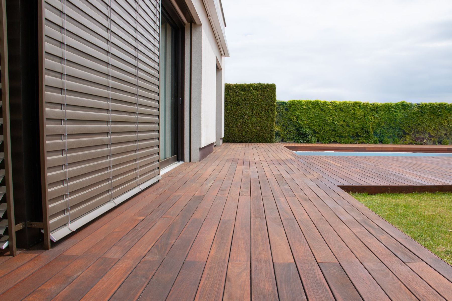 A wooden deck extends from a modern house toward a swimming pool, bordered by a green hedge and grass.