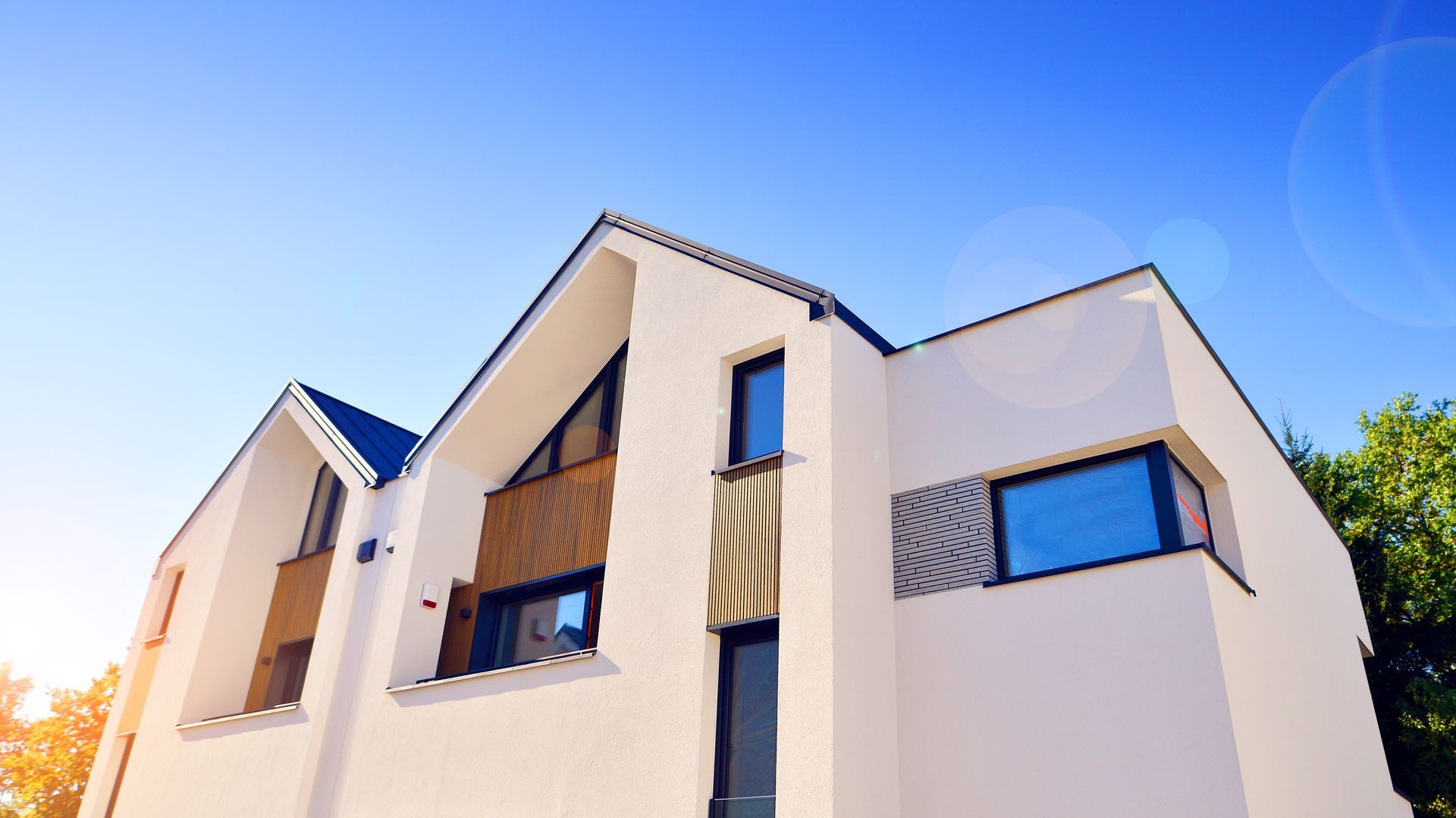 A low-angle view of a modern two-story house with white walls, wood accents, blue windows, and a clear blue sky background.