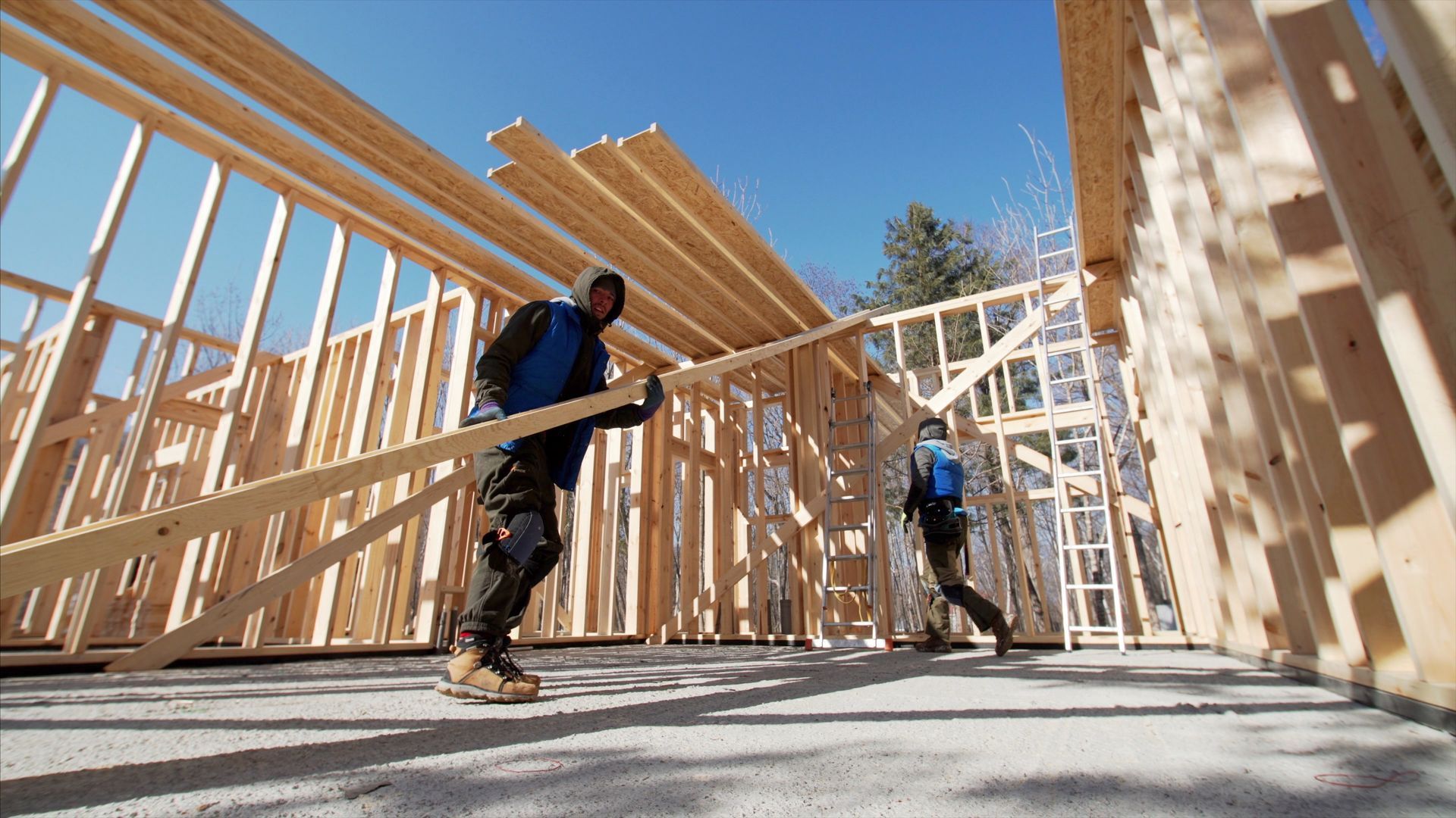 Two construction workers carry wooden beams inside the unfinished frame of a building under a bright blue sky.