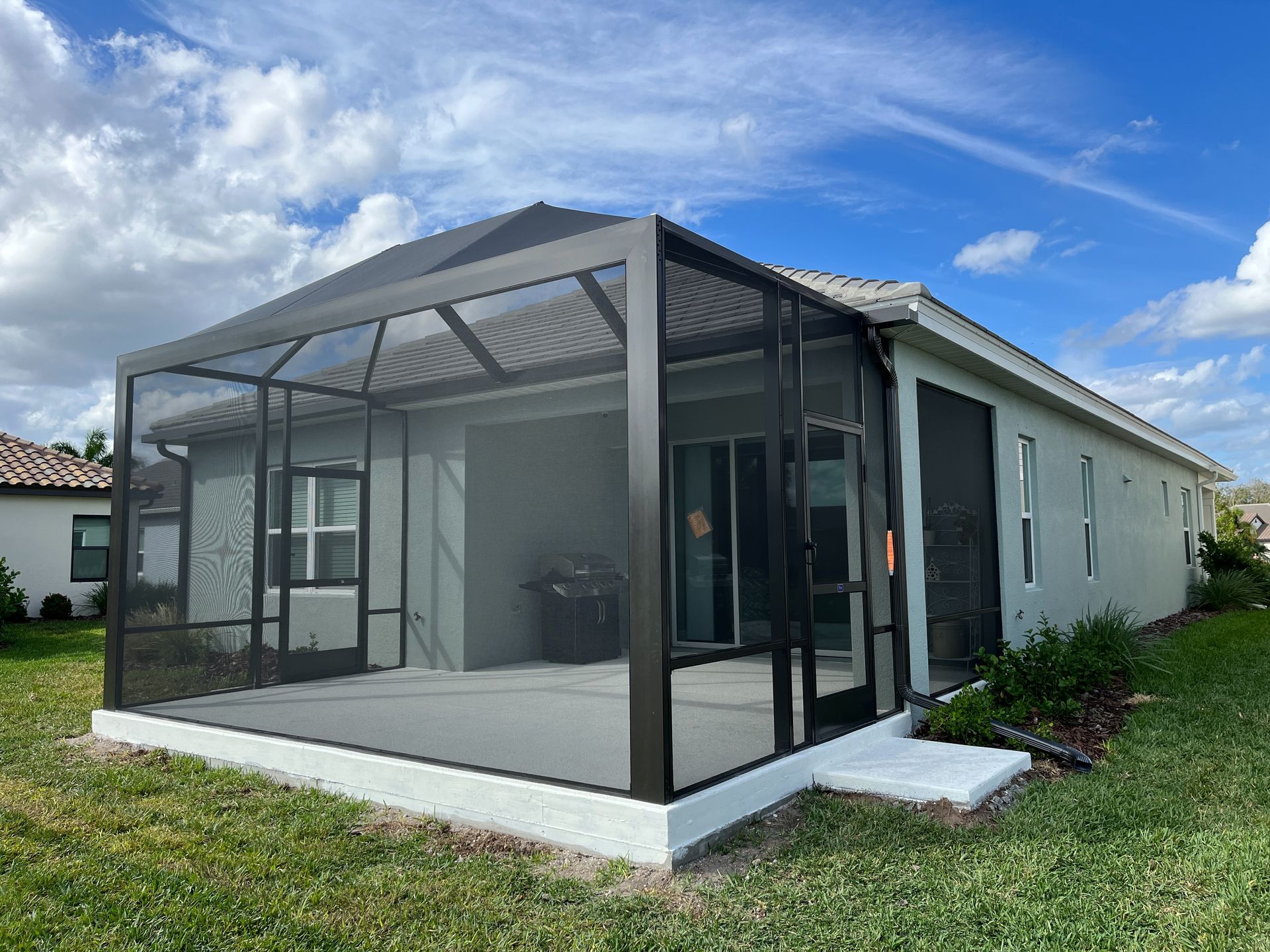 A back view of a light-colored house with a dark-framed screen-enclosed patio, set in a grassy yard under a blue sky.