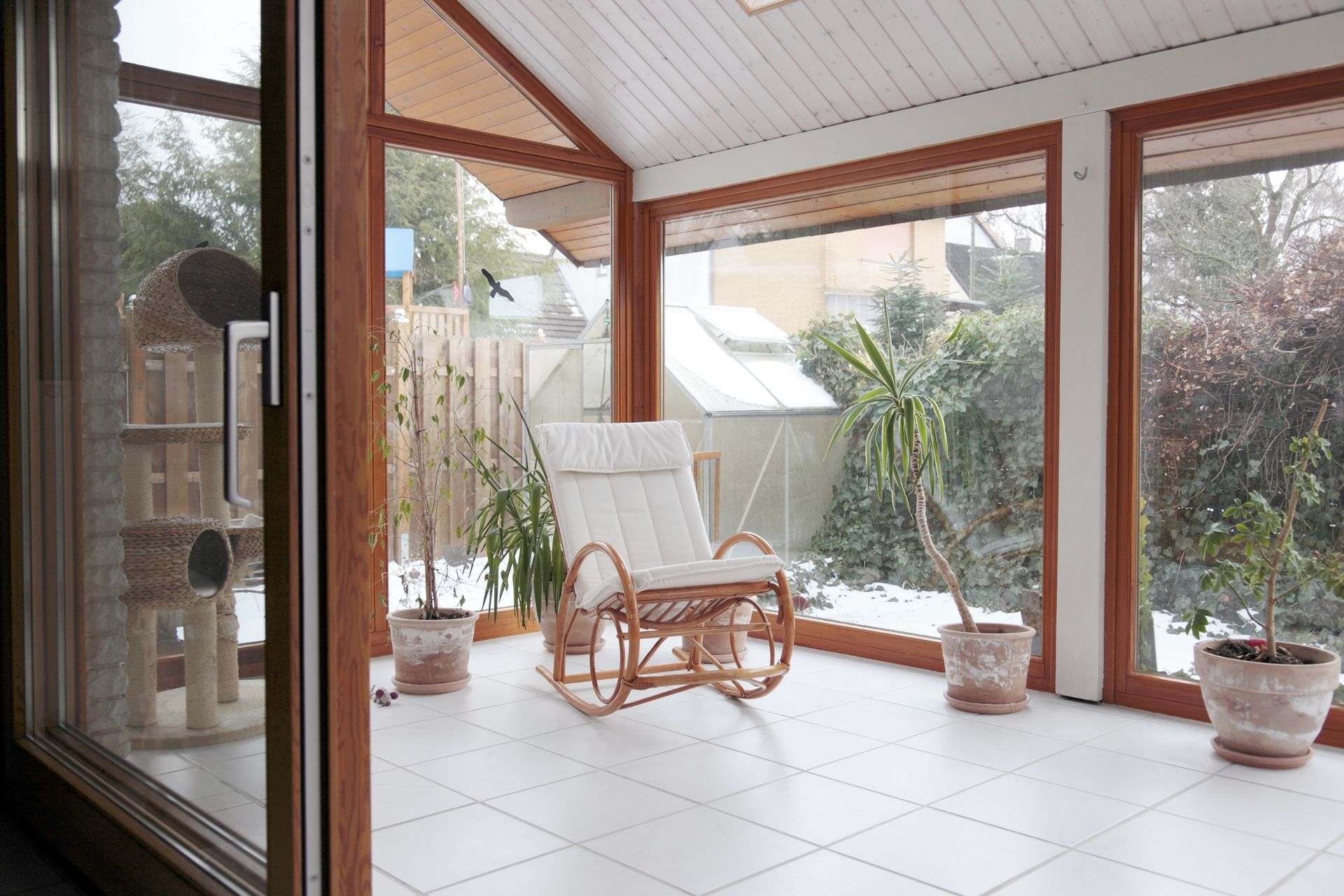 A sunroom with a rocking chair, potted plants, and a cat tree, looking out onto a snowy backyard.