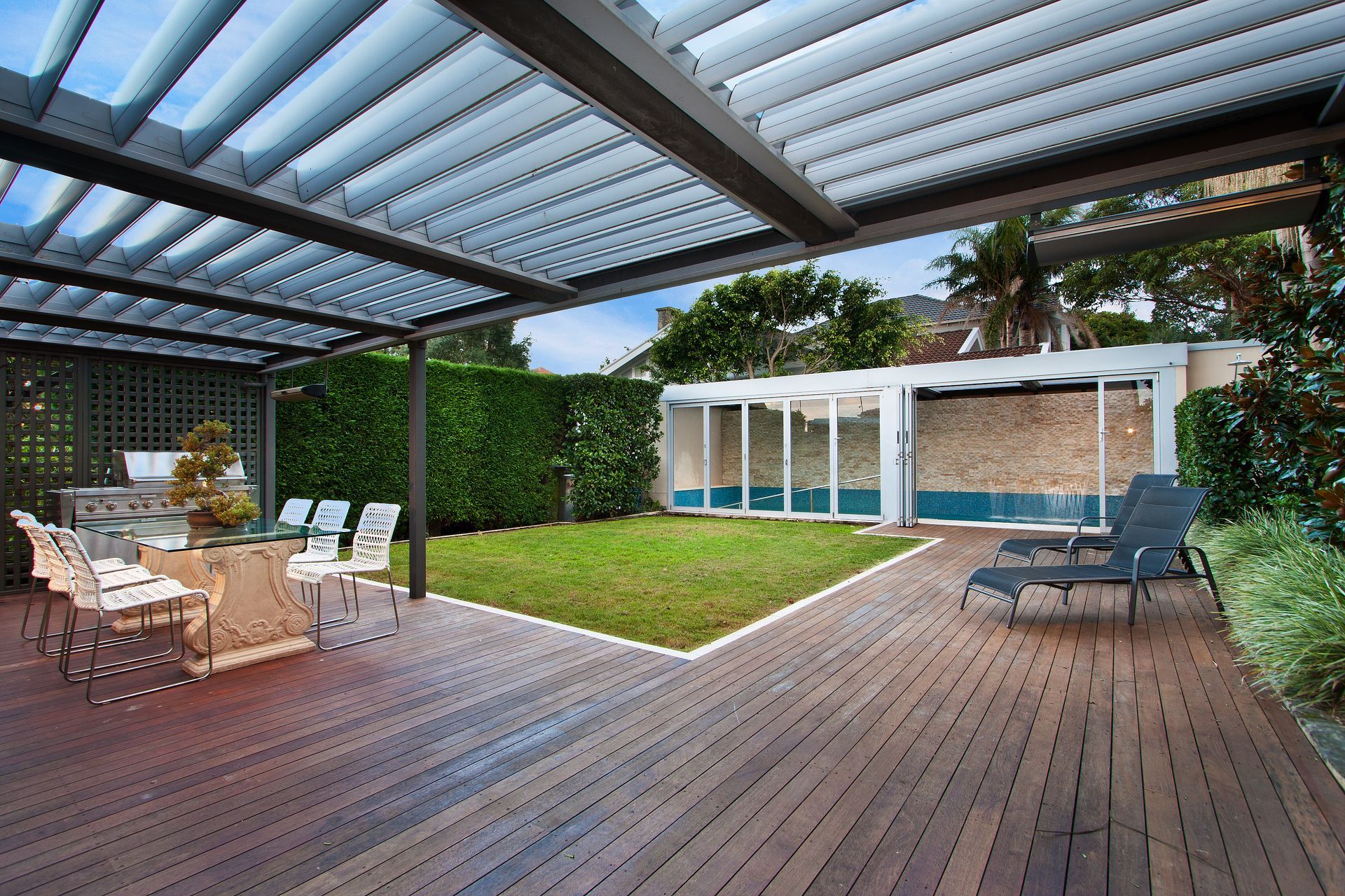 A wooden deck with an outdoor dining set under a pergola, a small lawn, and a pool house with glass doors.