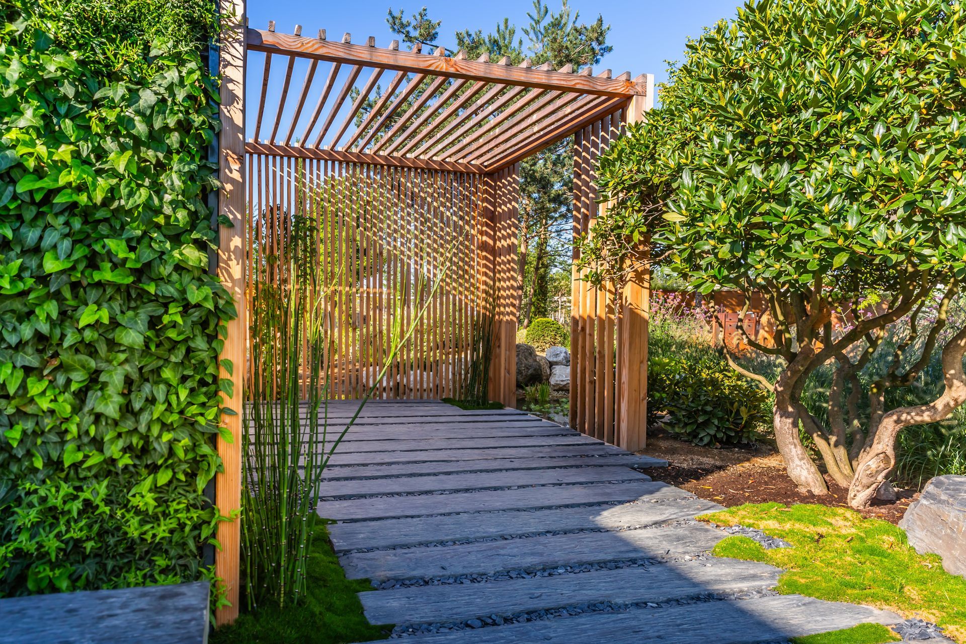 A wooden pergola and a slatted screen stand at the end of a stone walkway in a sunlit garden, flanked by lush greenery.