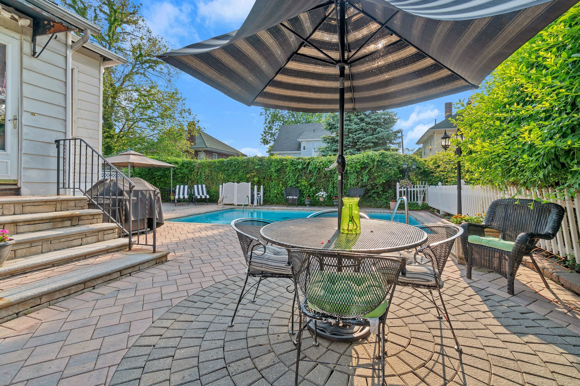 A patio with a round metal table, four chairs, and a striped umbrella next to a swimming pool and a house.
