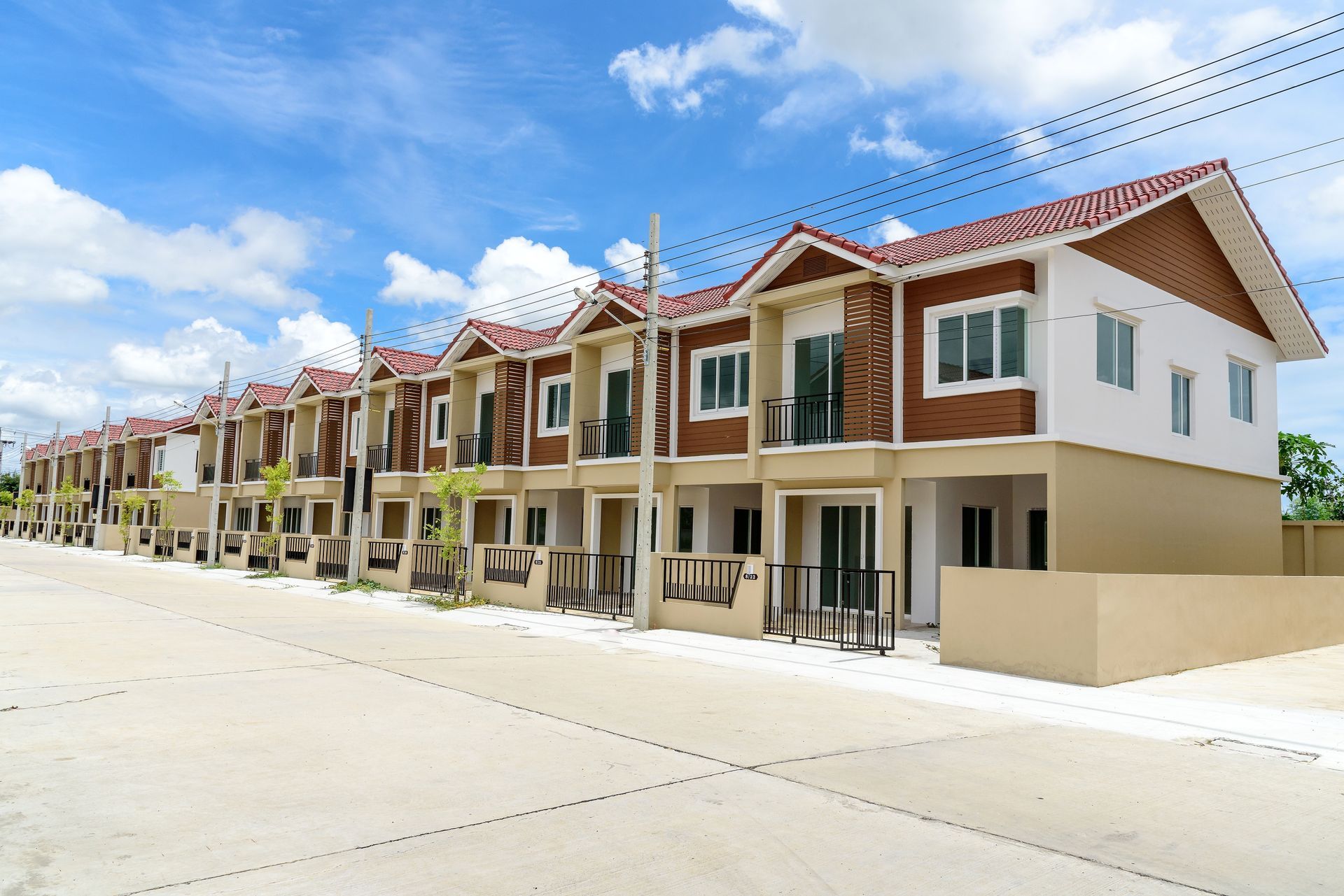 A row of newly built, identical two-story townhouses with tan and brown exteriors under a bright, cloudy blue sky.
