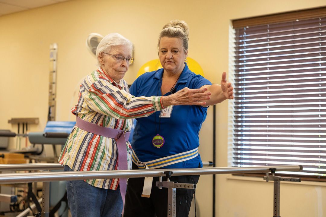 A woman is helping an elderly woman walk on parallel bars.