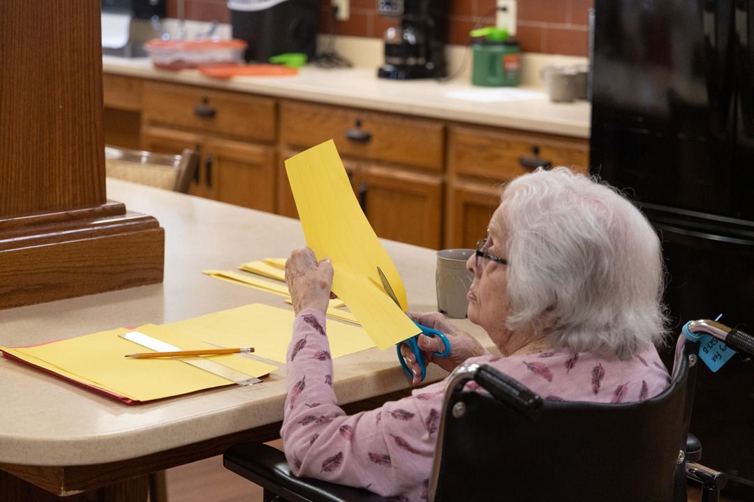 An elderly woman in a wheelchair is cutting a piece of paper with scissors.