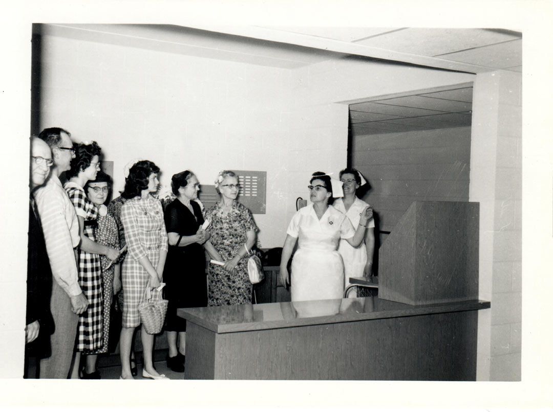 A black and white photo of a group of people standing around a desk