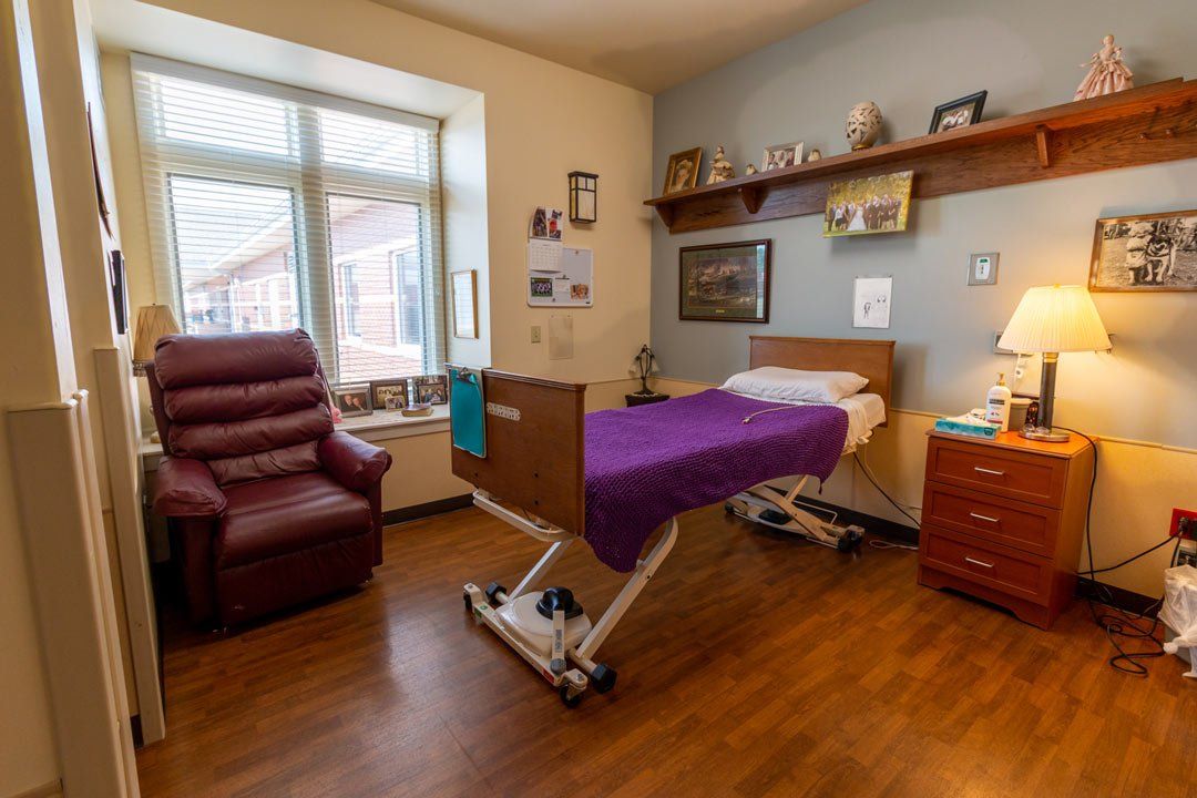 A hospital room with a bed , chair , nightstand and window.