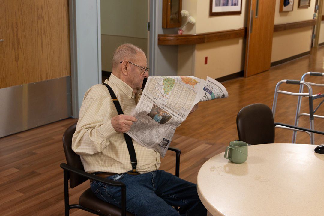 An elderly man is sitting at a table reading a newspaper.