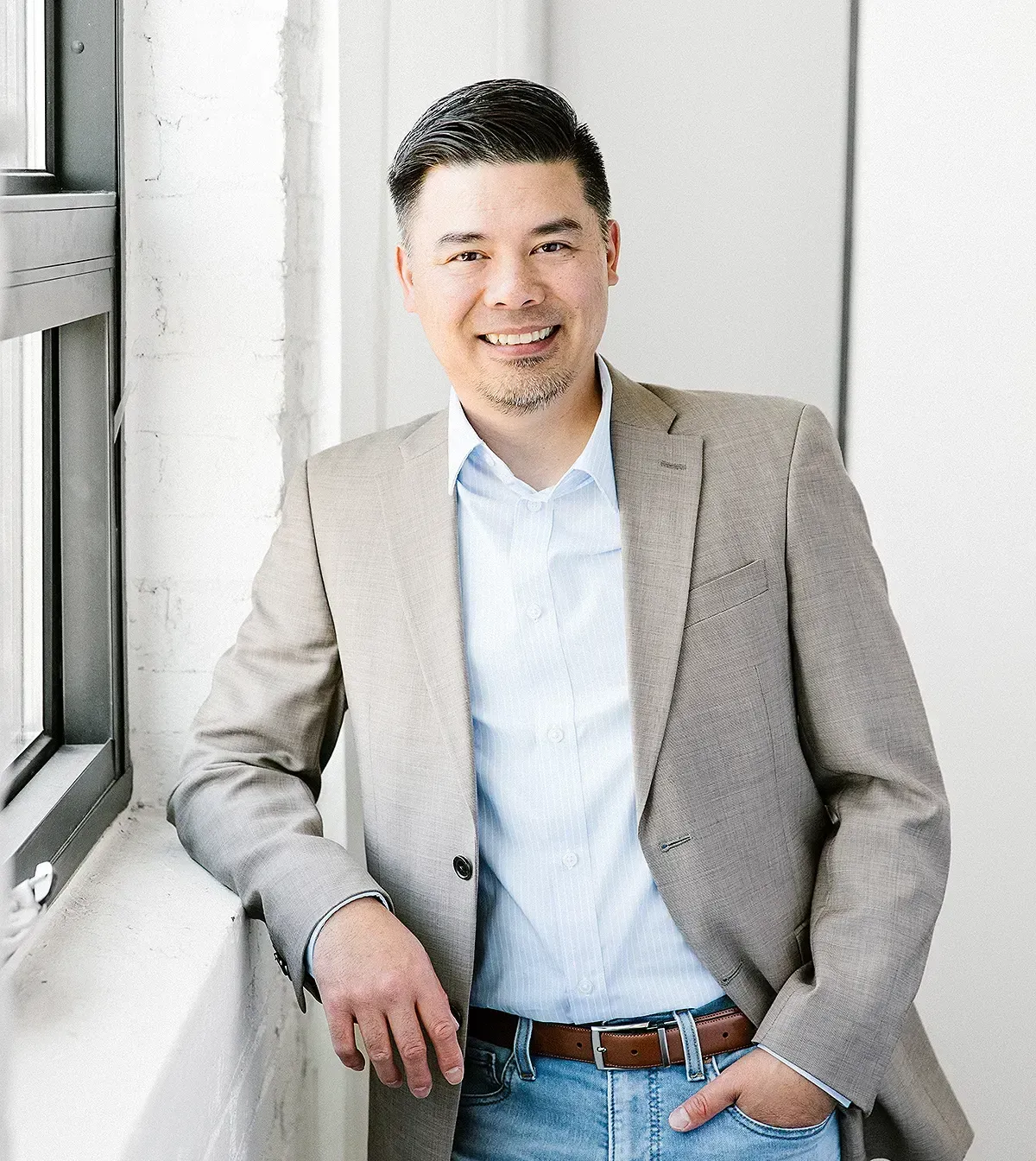 A man in a suit and jeans is leaning against a window.
