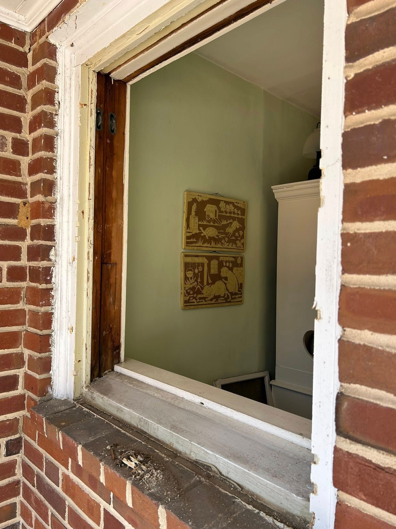 Window frame view of an interior room: green walls, two framed artworks, white cabinet.