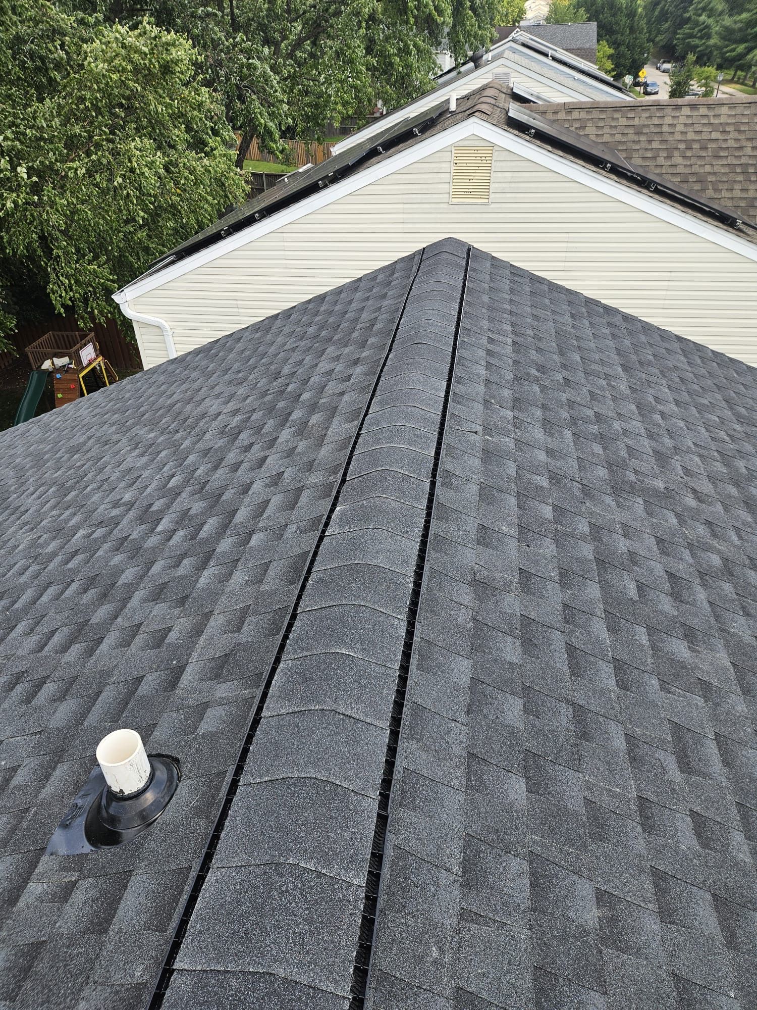 Dark gray shingle roof with a central seam; a vent pipe and part of another house with beige siding and a dark roof.