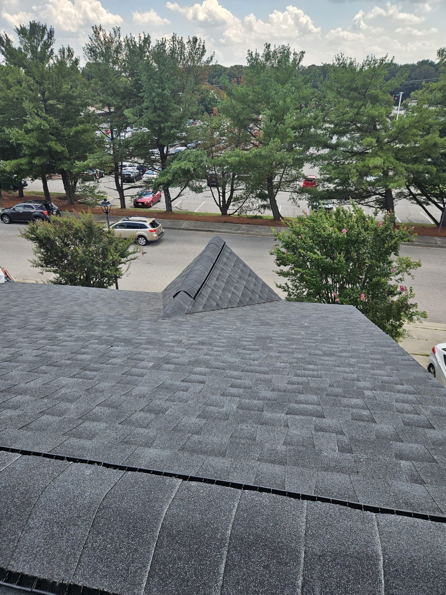 View from a dark shingled roof with a peak, overlooking a street with parked cars and trees.