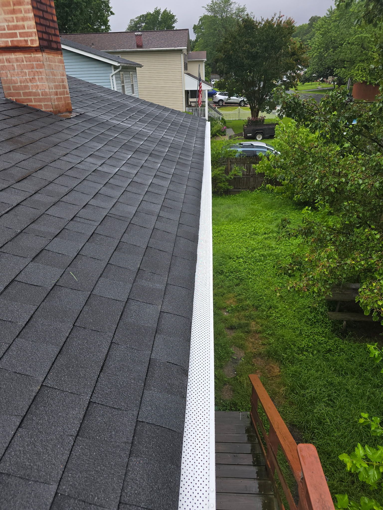Black asphalt shingle roof with white gutter and green vegetation alongside wooden stairs.