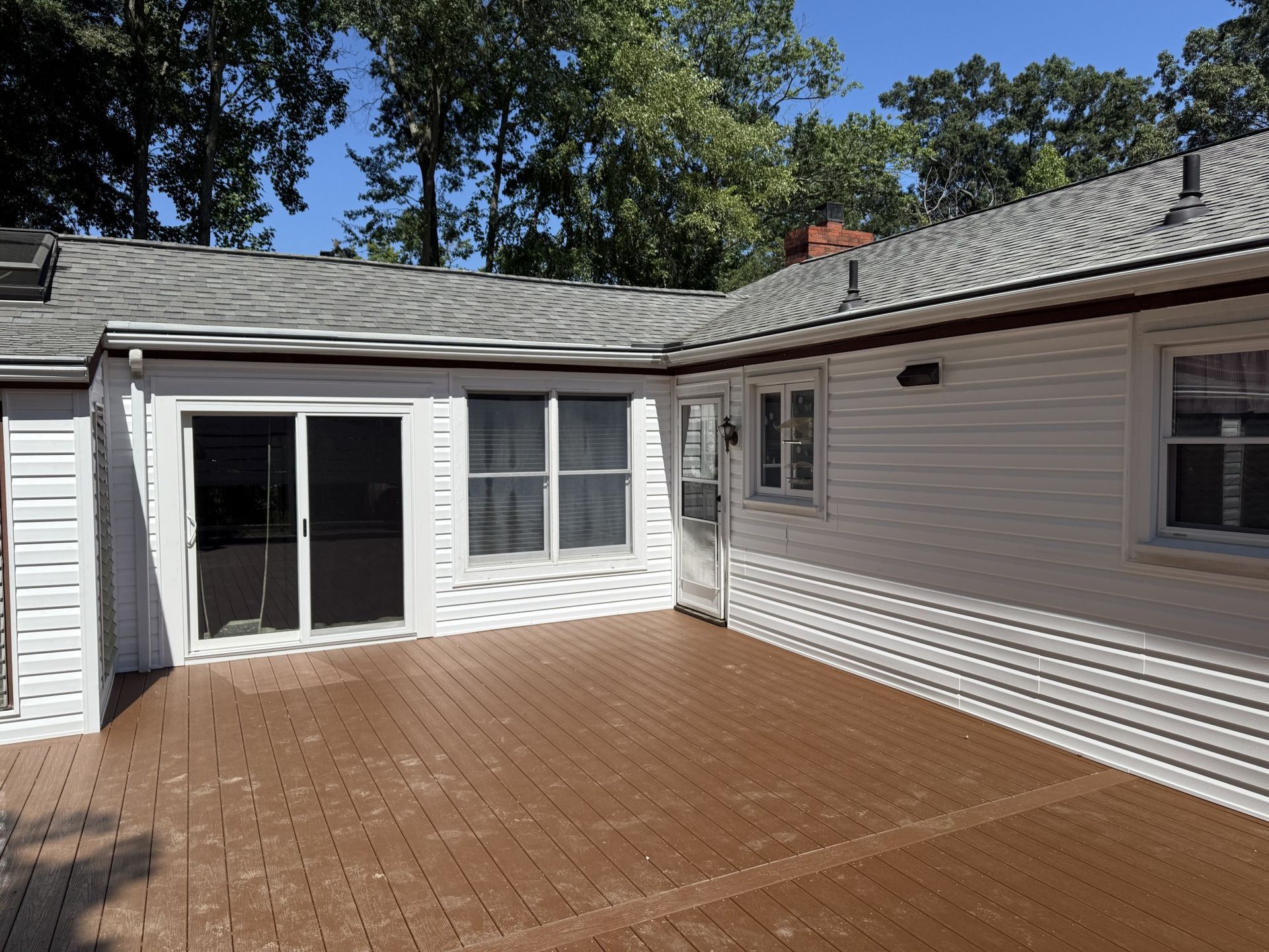 Brown wooden deck attached to a white house with a sliding glass door and windows.