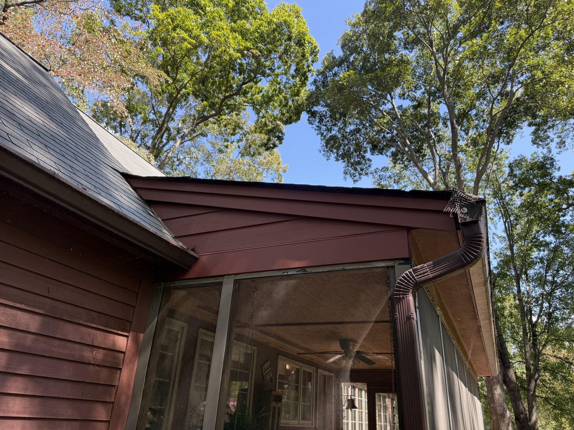 Brown house with a screened porch and dark brown roof against a backdrop of trees and blue sky.