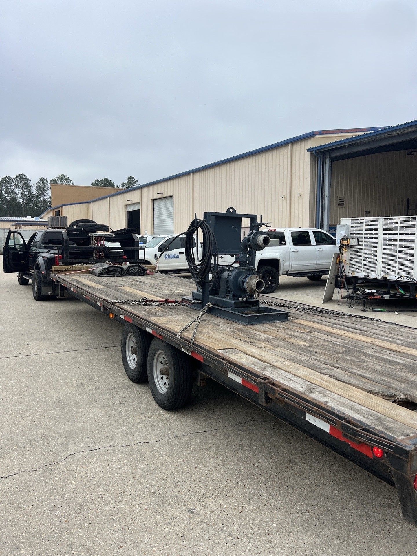 Flatbed trailer loaded with metal equipment parked in an industrial lot near parked vehicles.