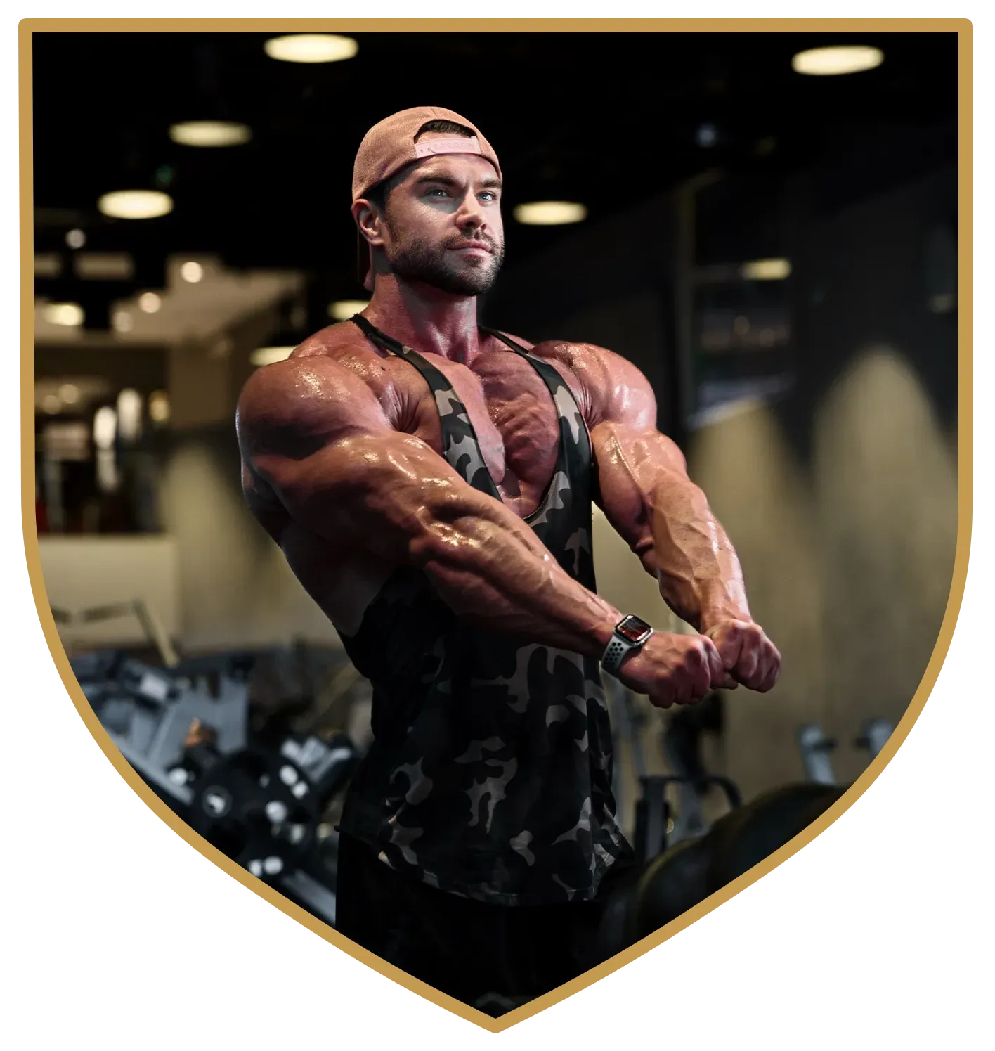 Muscular individual in a camouflage tank top and cap posing in a gym, highlighting arm and shoulder muscle definition.