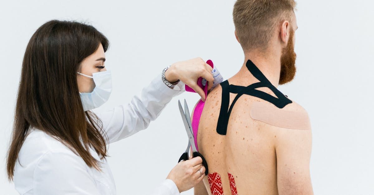 Person with red beard has tape applied to his back while medical worker cuts excess tape with scissors.