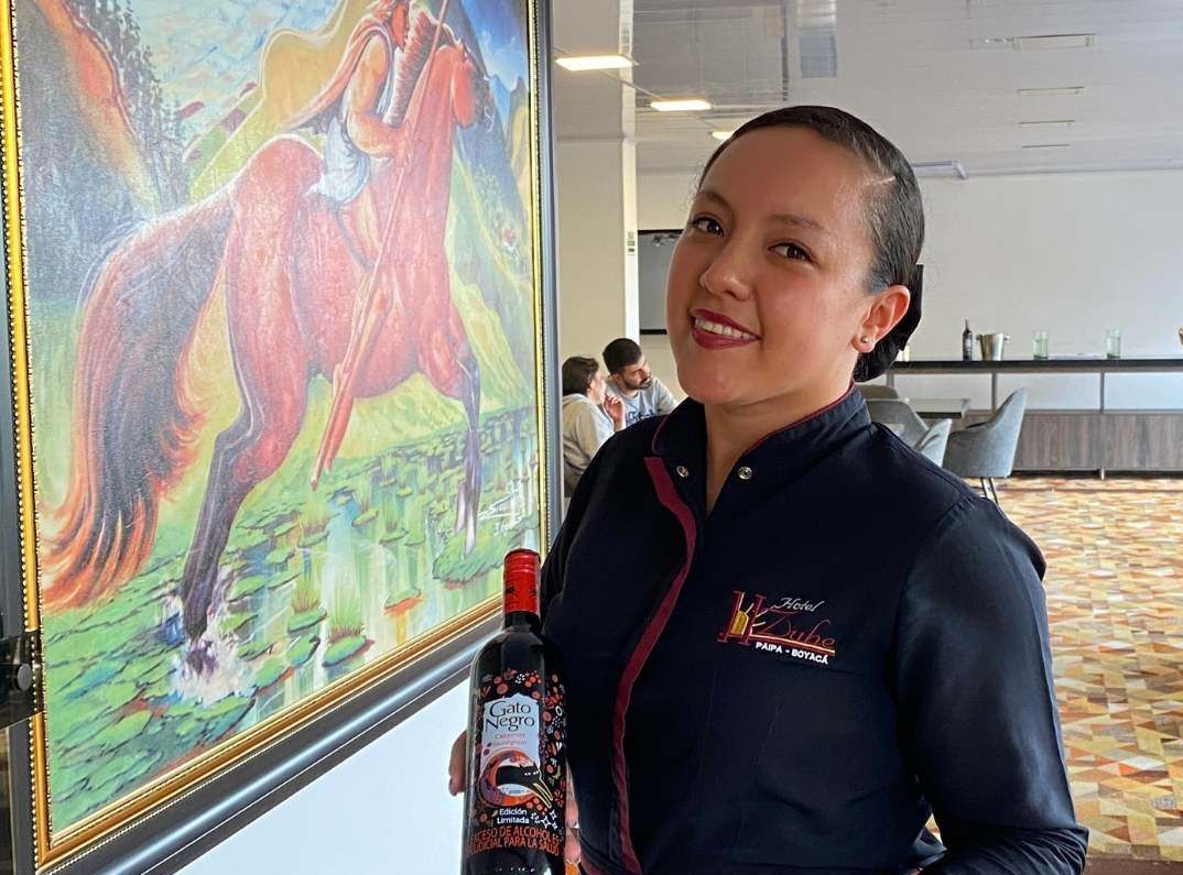 Woman holding a wine bottle next to a colorful horse painting. She smiles in a restaurant setting.