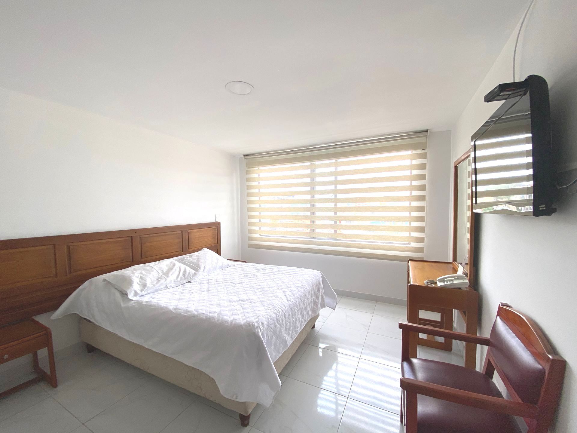 Bedroom with a large bed, window with blinds, TV on the wall, and a wooden chair.