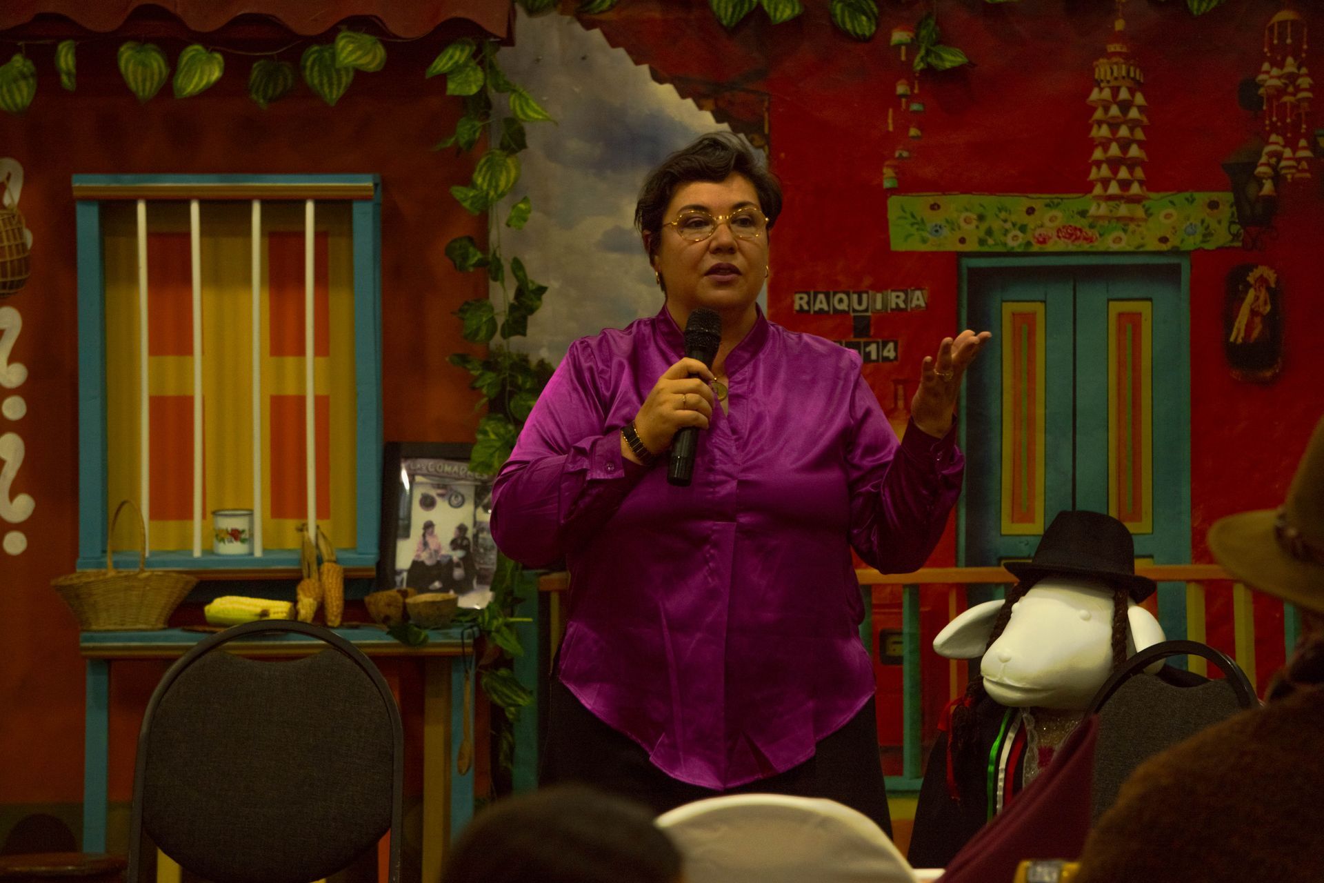 Woman in purple shirt speaking into a microphone, gesturing. Colorful background with a sheep in a hat.