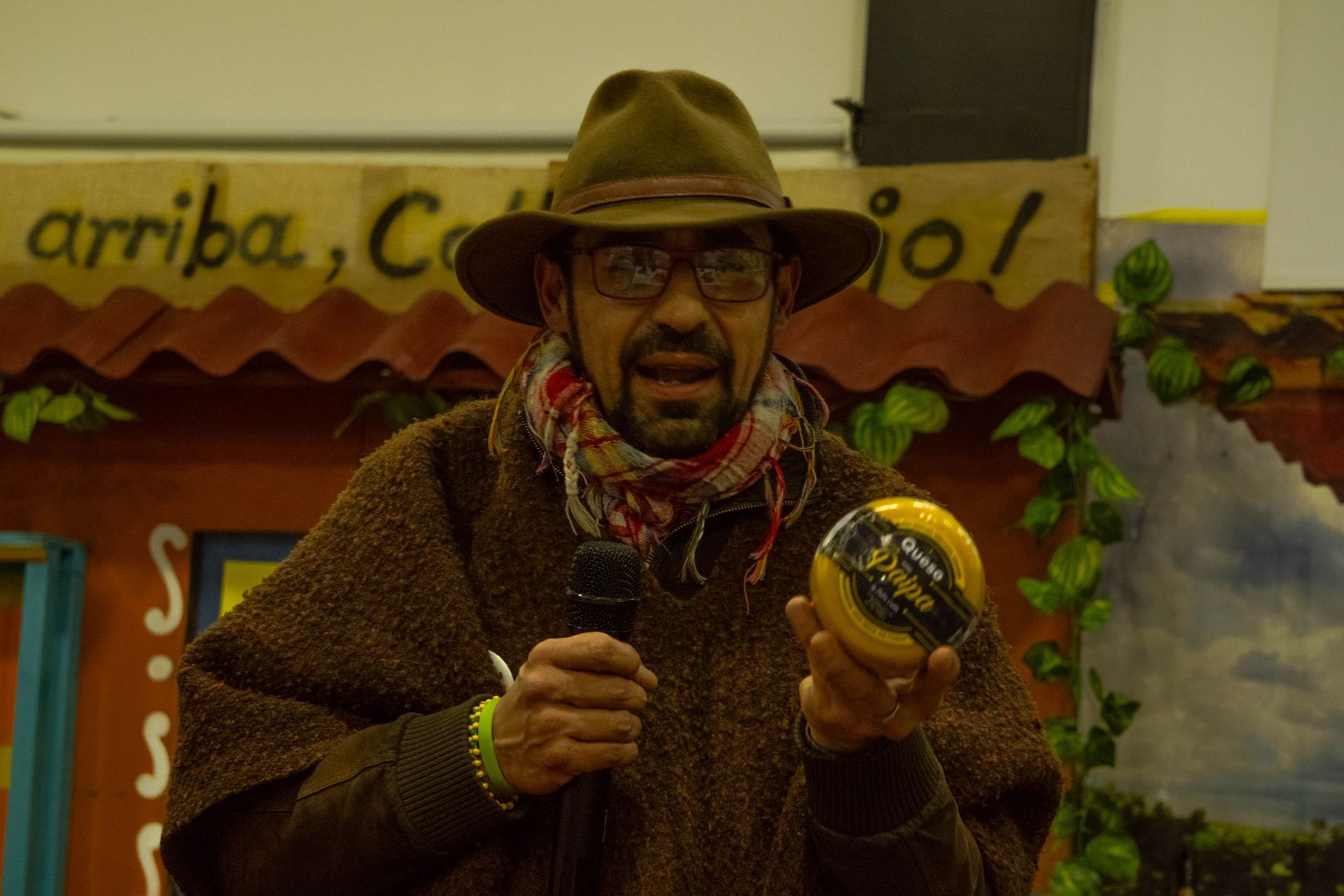 Man in hat and poncho holding a round cheese, speaking into a microphone, colorful background.